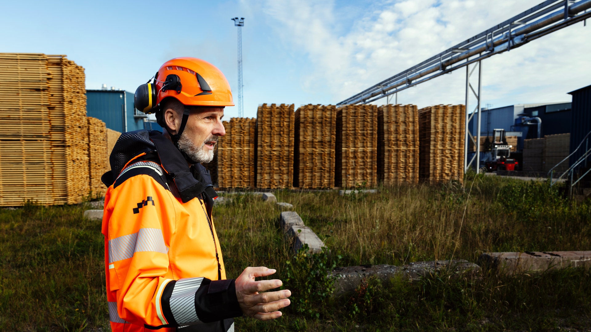 En man i orange skyddskläder och hjälm står vid ett sågverk med staplade timmerpaket i bakgrunden.