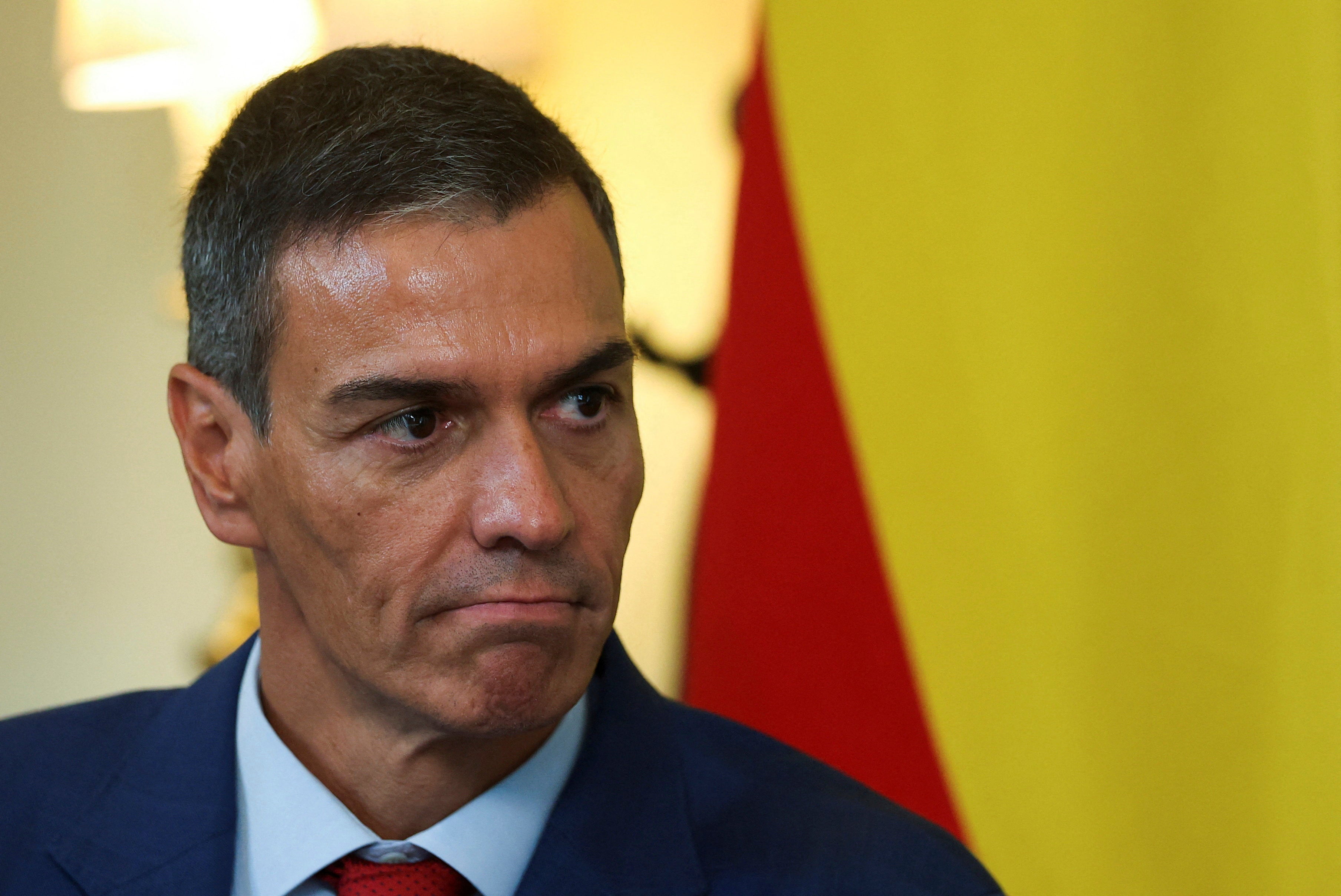 Porträtt av Pedro Sánchez i mörk kostym och röd slips med allvarlig blick. Spaniens flagga syns i bakgrunden.