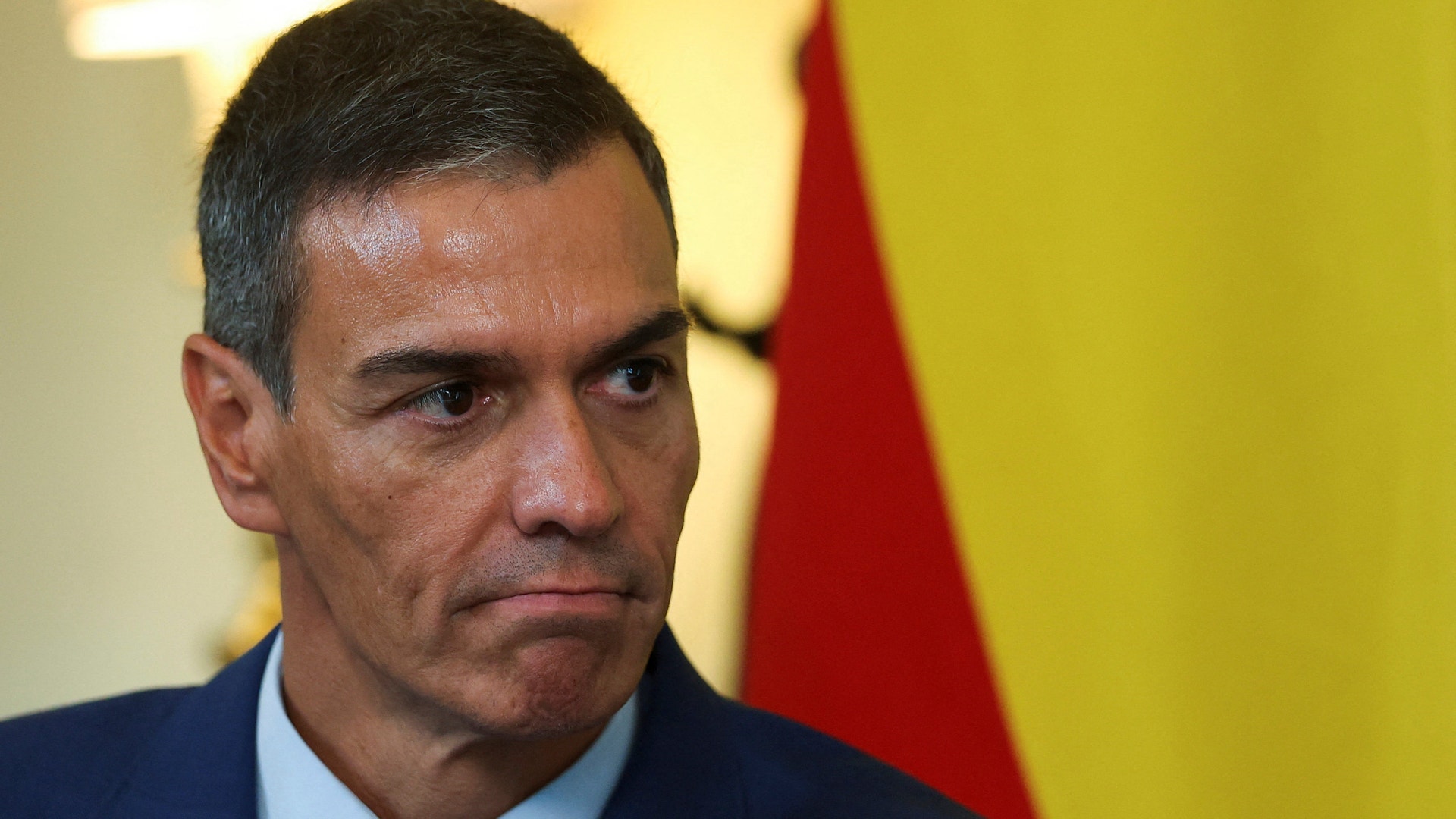 Porträtt av Pedro Sánchez i mörk kostym och röd slips med allvarlig blick. Spaniens flagga syns i bakgrunden.