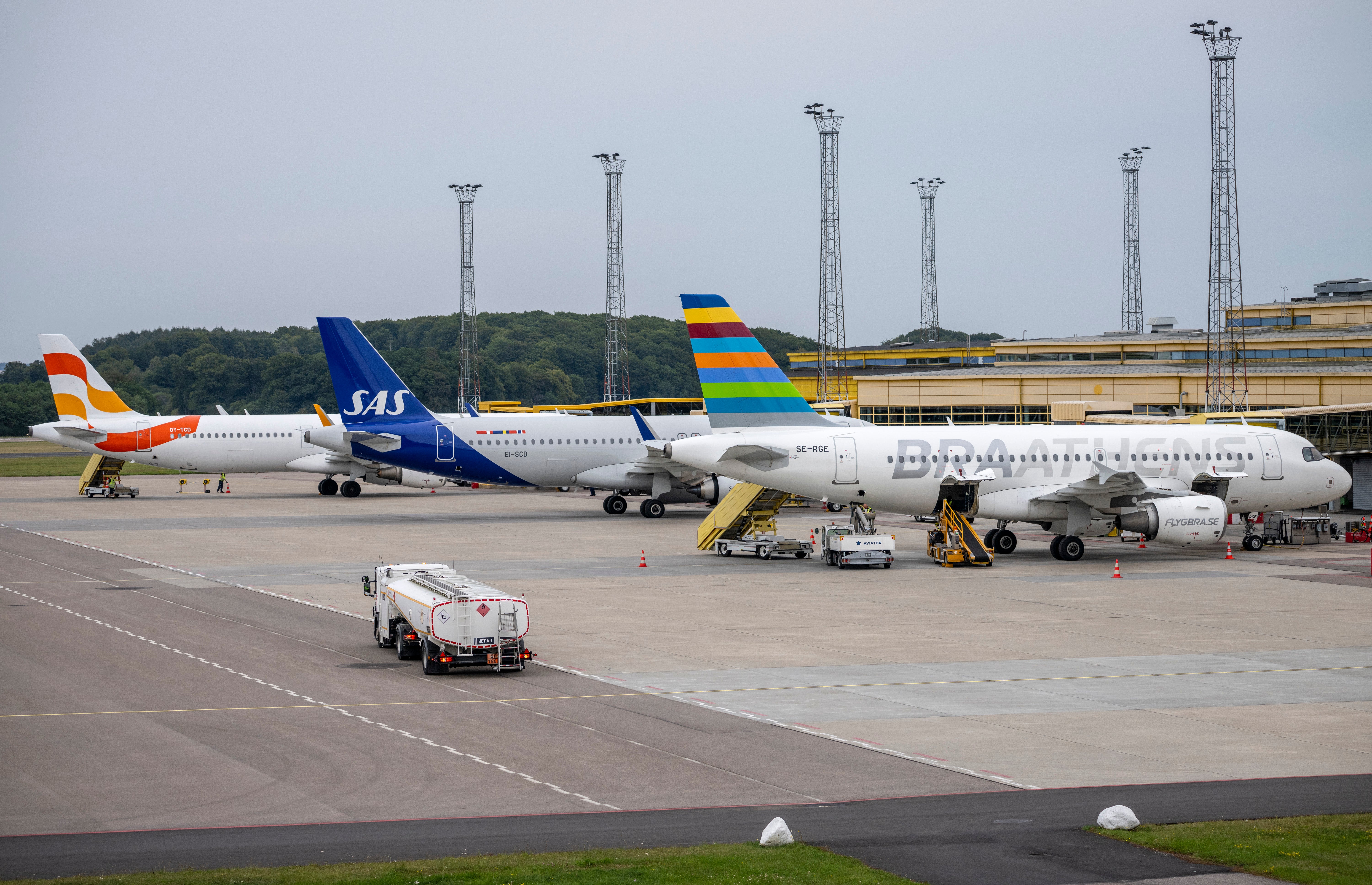 Flera passagerarflygplan parkerade vid en flygplats terminal med markservicefordon och luftfartstorn i bakgrunden.