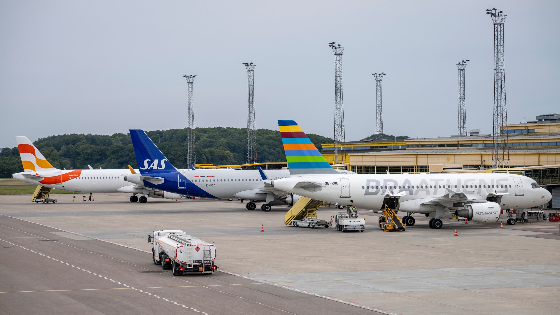 Flera passagerarflygplan parkerade vid en flygplats terminal med markservicefordon och luftfartstorn i bakgrunden.