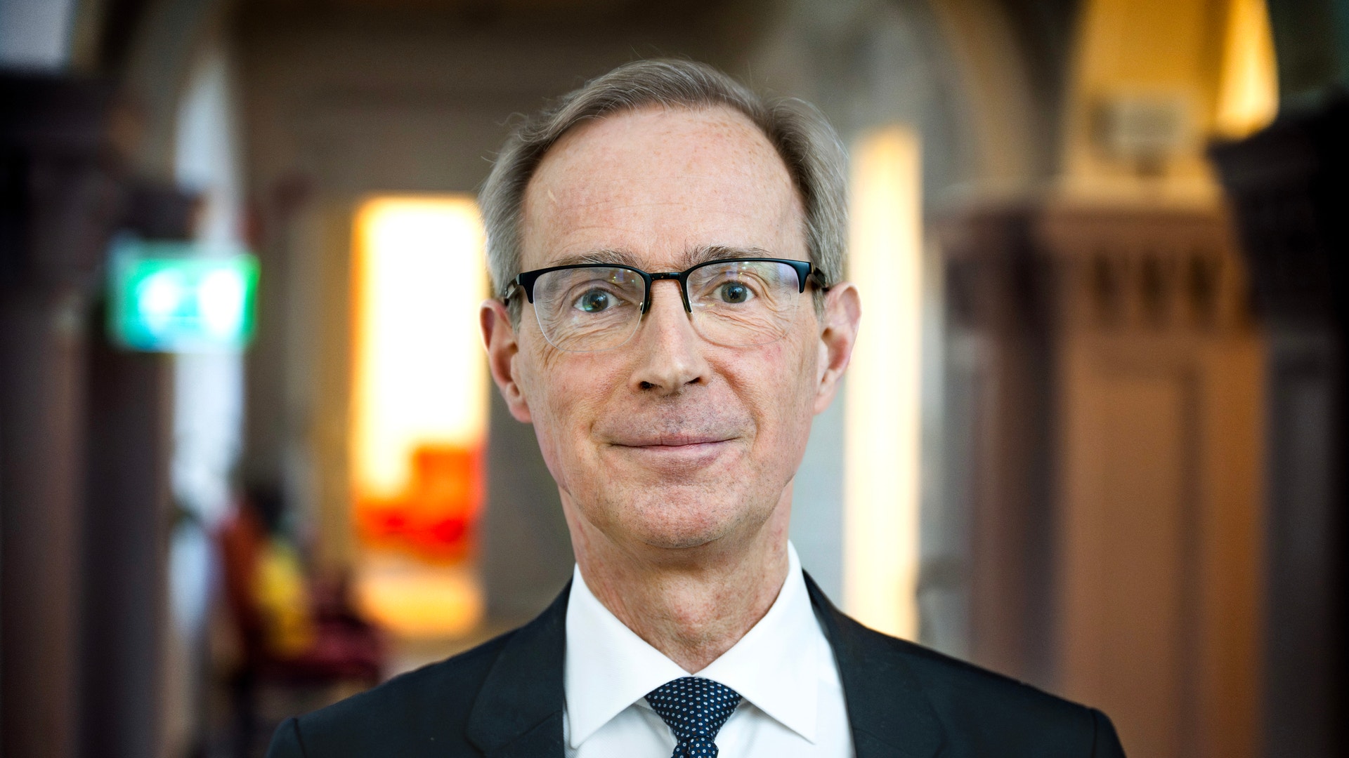 Porträttfoto av en man i glasögon och kostym med blå slips, fotograferad inomhus.