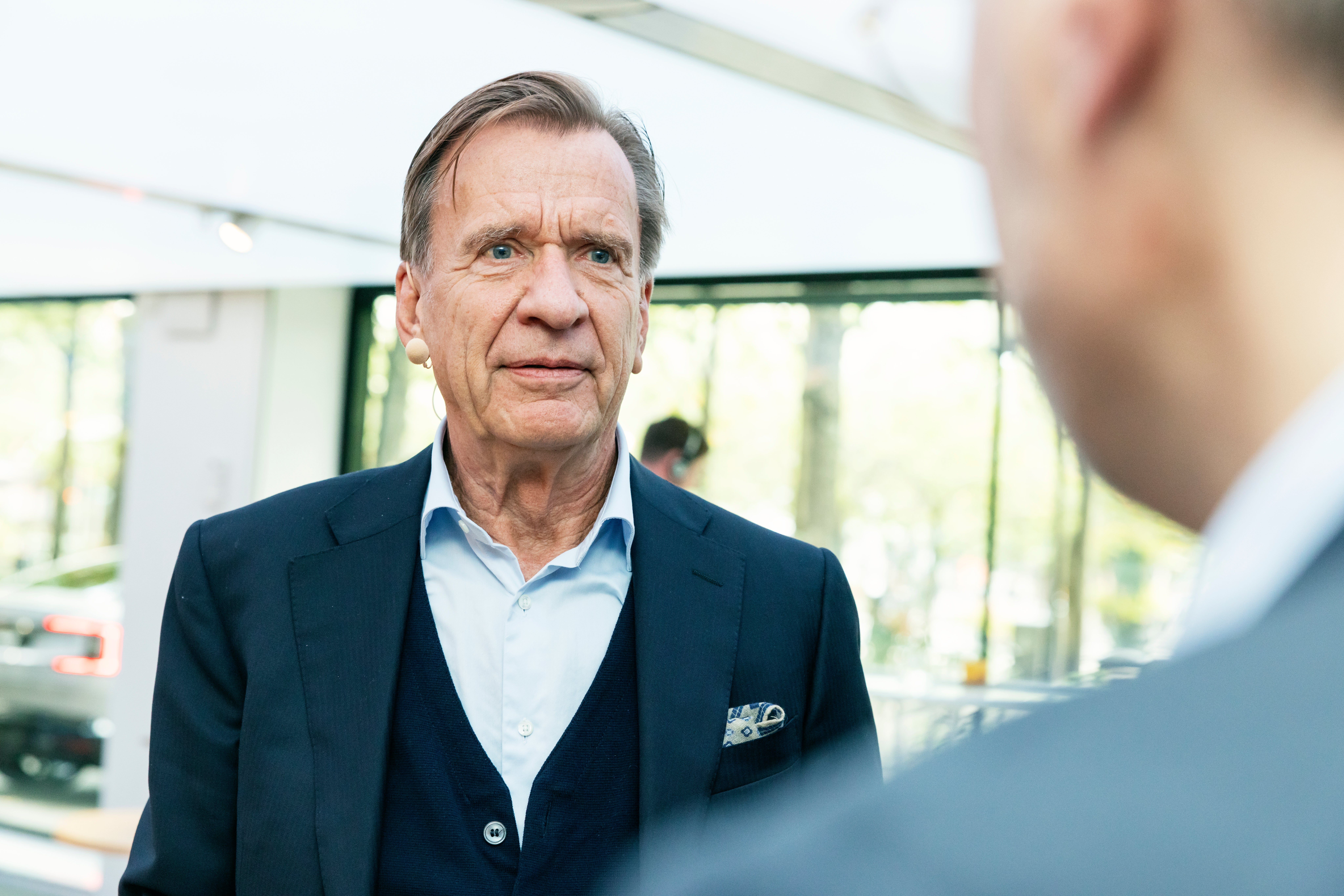 En äldre man i kavaj pratar med en person som syns i förgrunden.