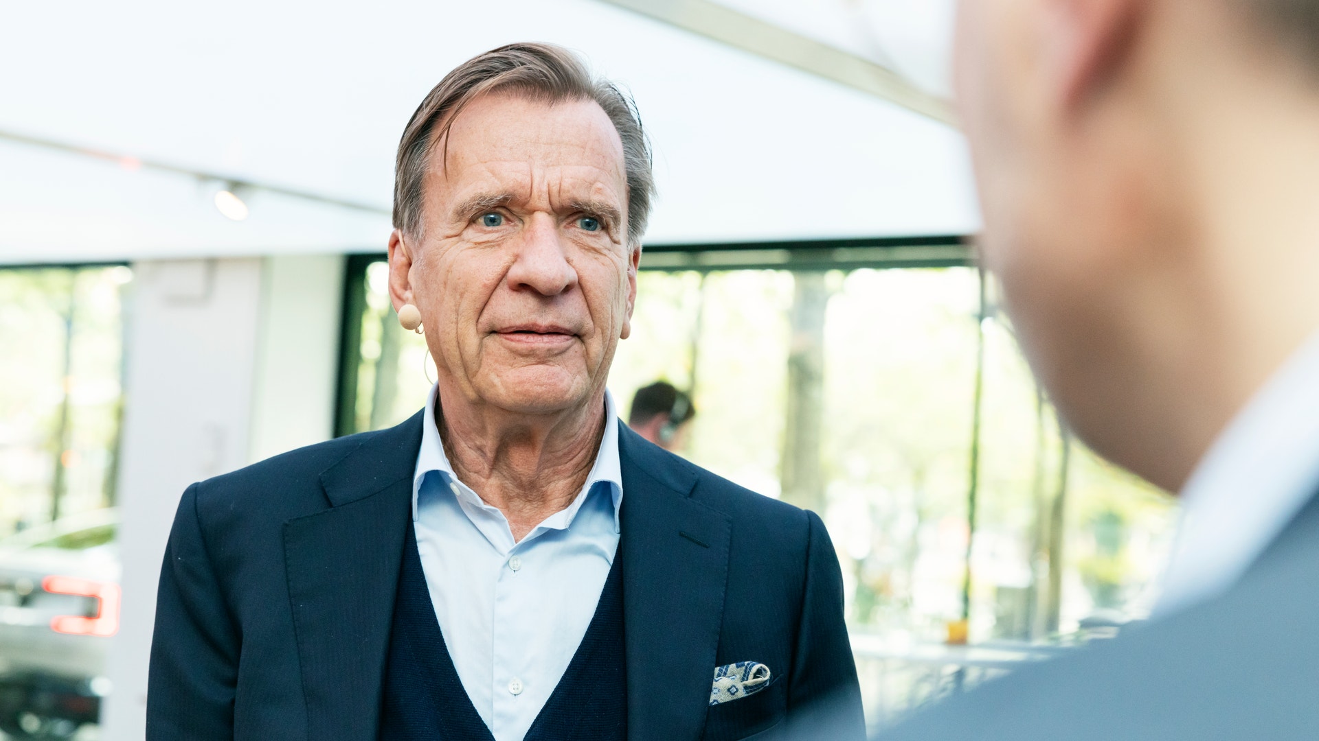 En äldre man i kavaj pratar med en person som syns i förgrunden.