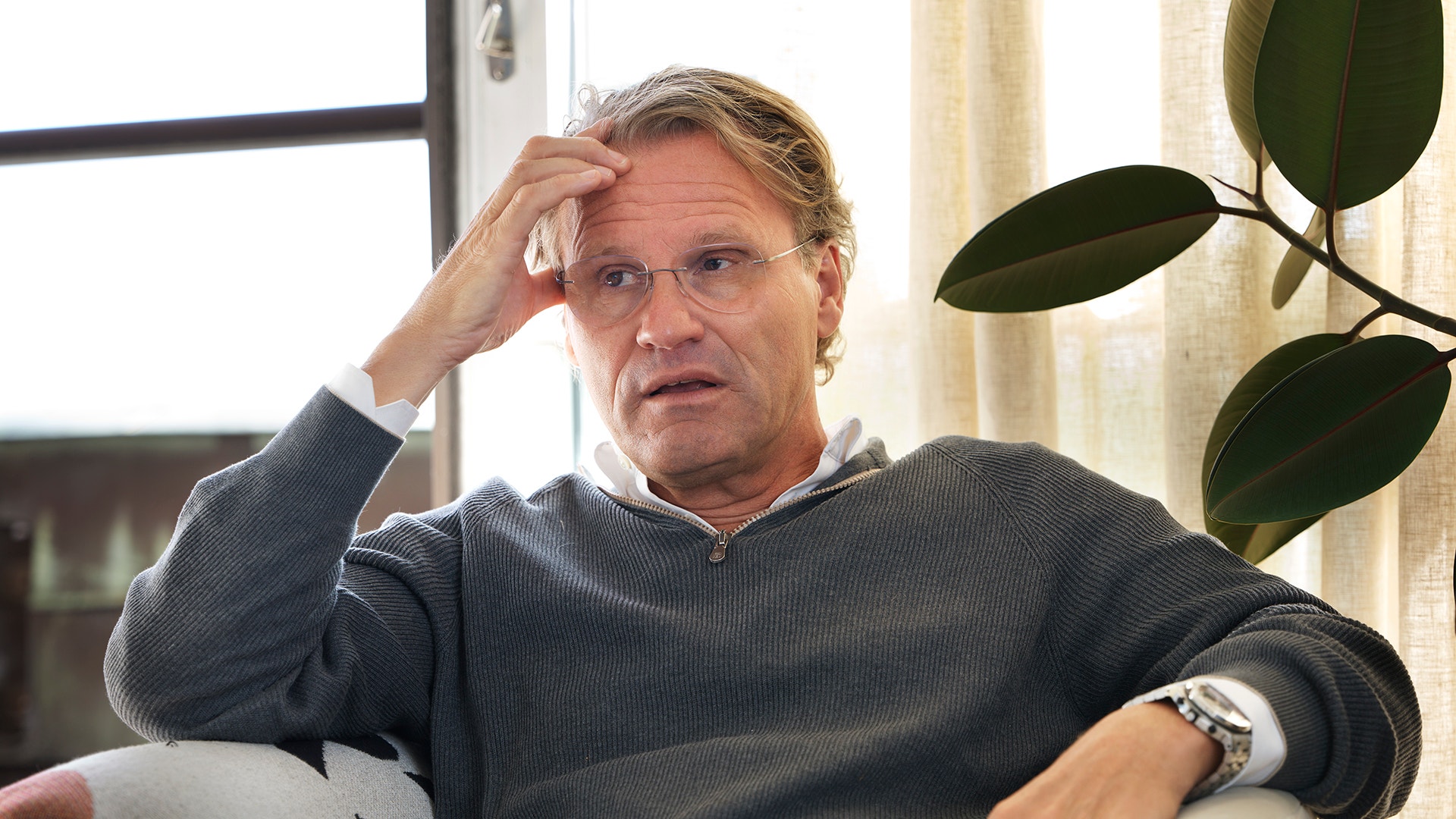 En man sitter i en soffa och pratar, med handen vid huvudet.