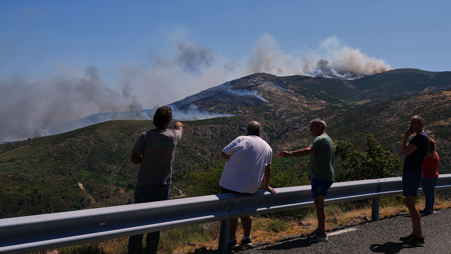 Flera personer står vid en väg och observerar en skogsbrand på en bergssluttning med rök och lågor synliga.