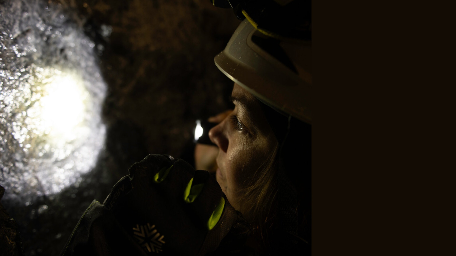 En kvinna med hjälm och pannlampa tittar mot en upplyst bergvägg i en mörk gruva.