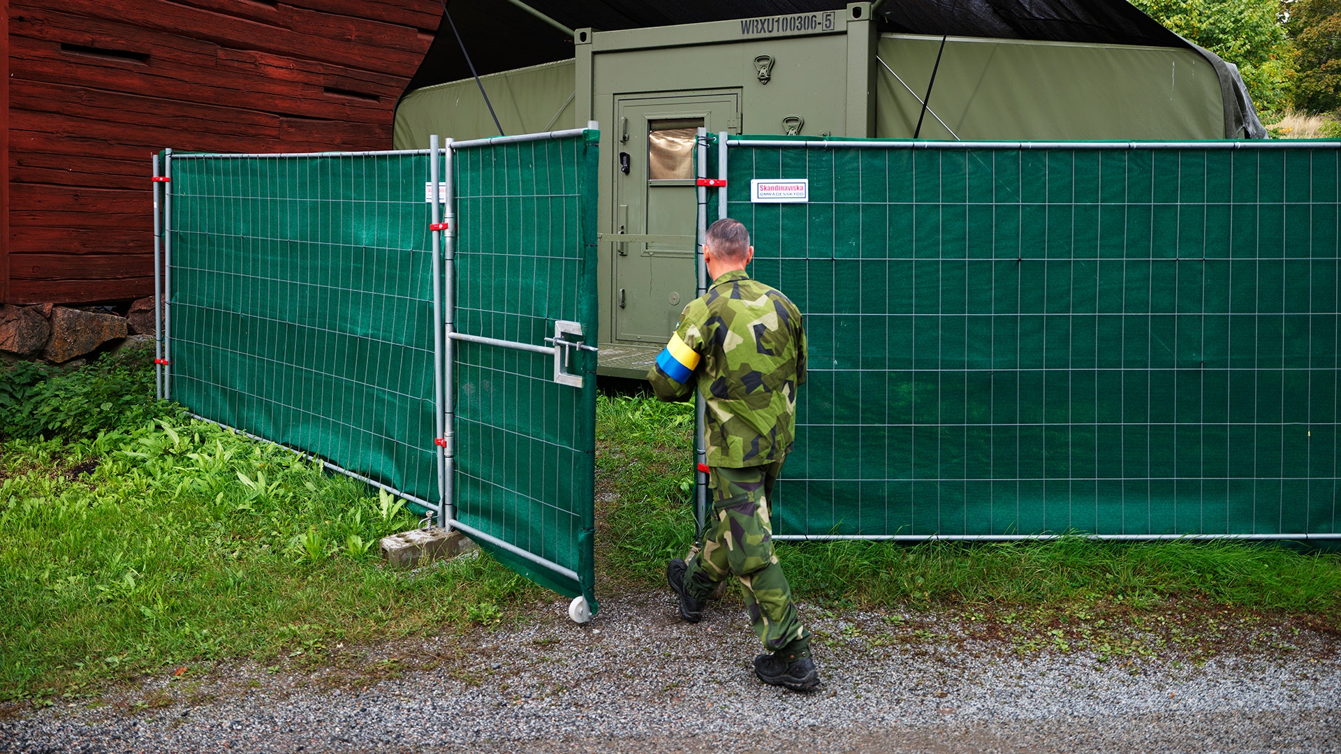 En soldat i kamouflageuniform går genom en grön grindöppning in till ett militärt fältläger med en container och en röd lada i bakgrunden.