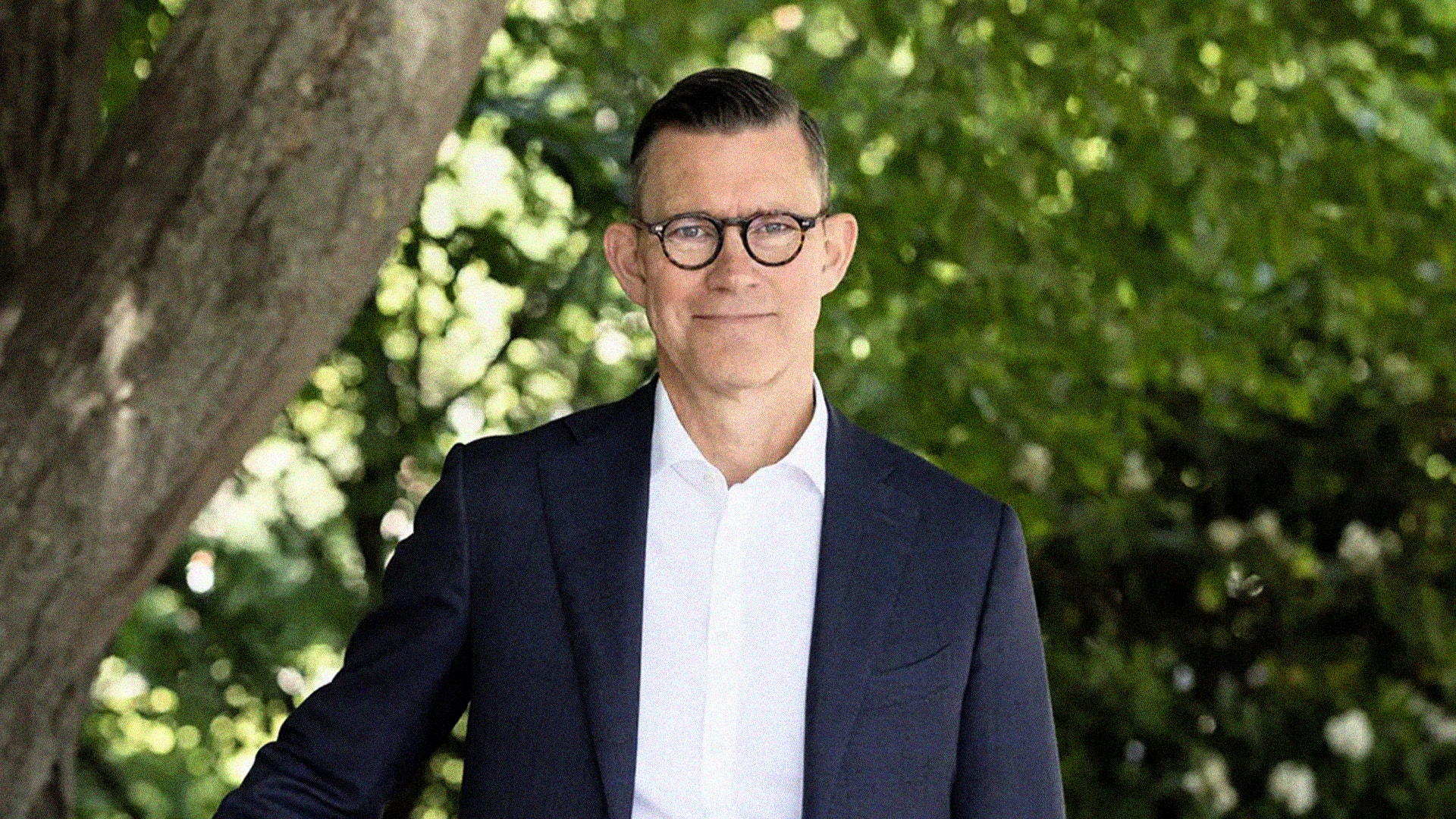 En man i kostym utan slips leende utomhus framför grönska.