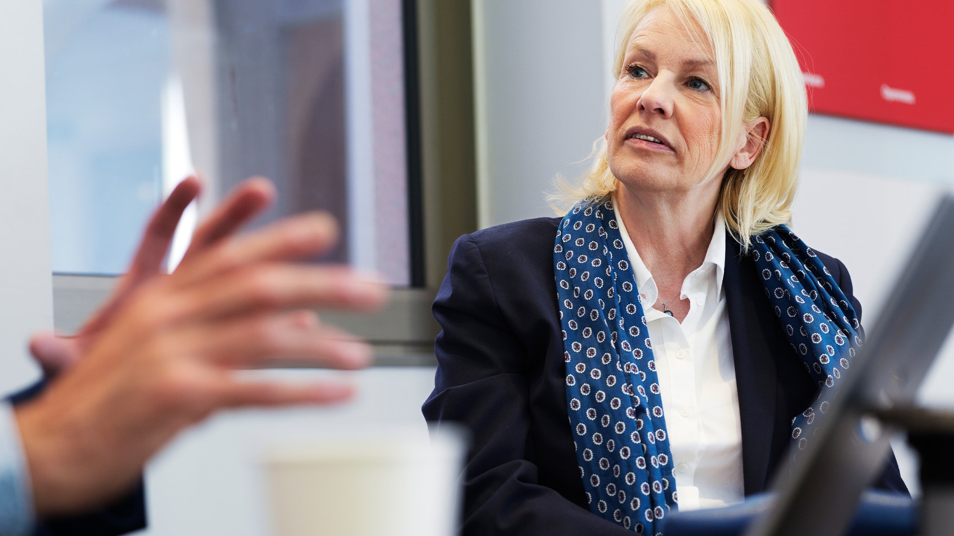 Kvinna i mörkblå kavaj och blå mönstrad scarf presenterar eller talar under ett möte.