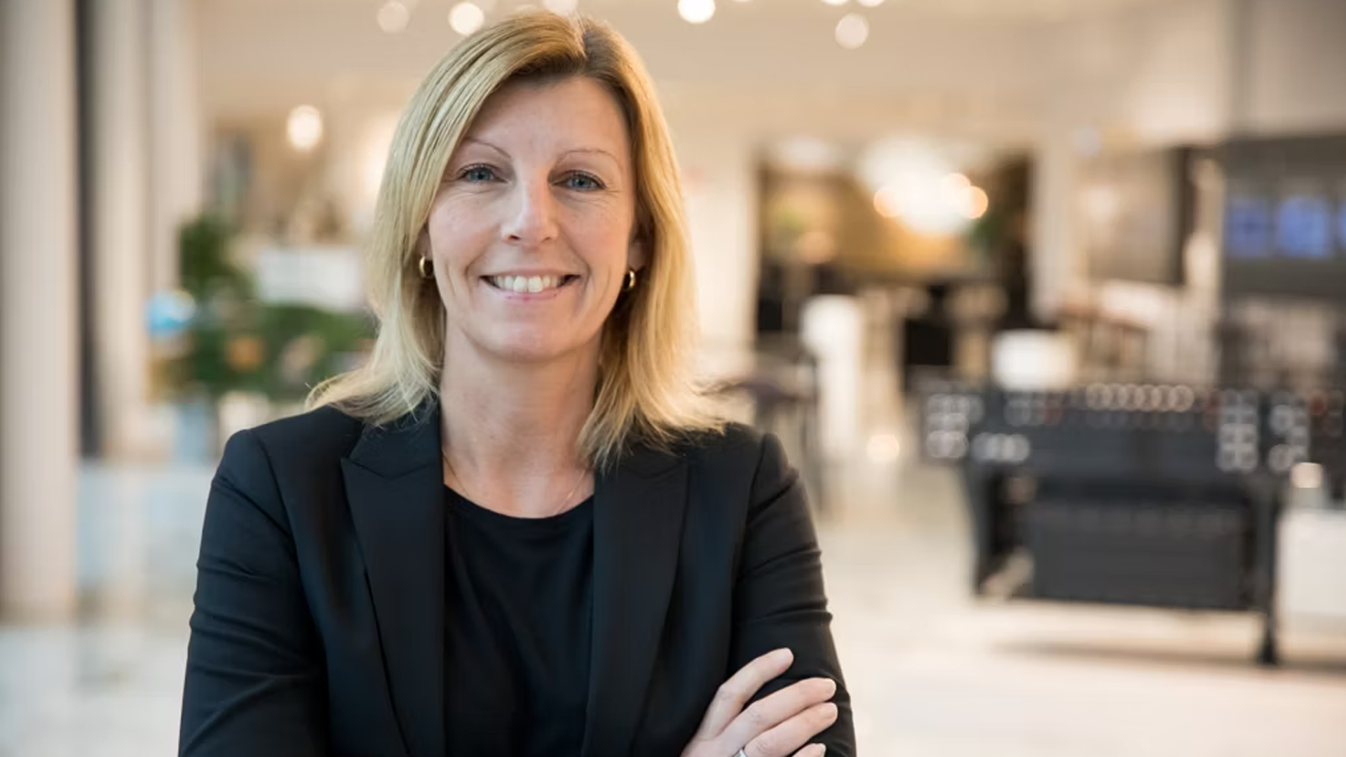 Porträttfoto av en kvinna i mörkblå blazer som står i en modern kontorsmiljö och ler mot kameran.