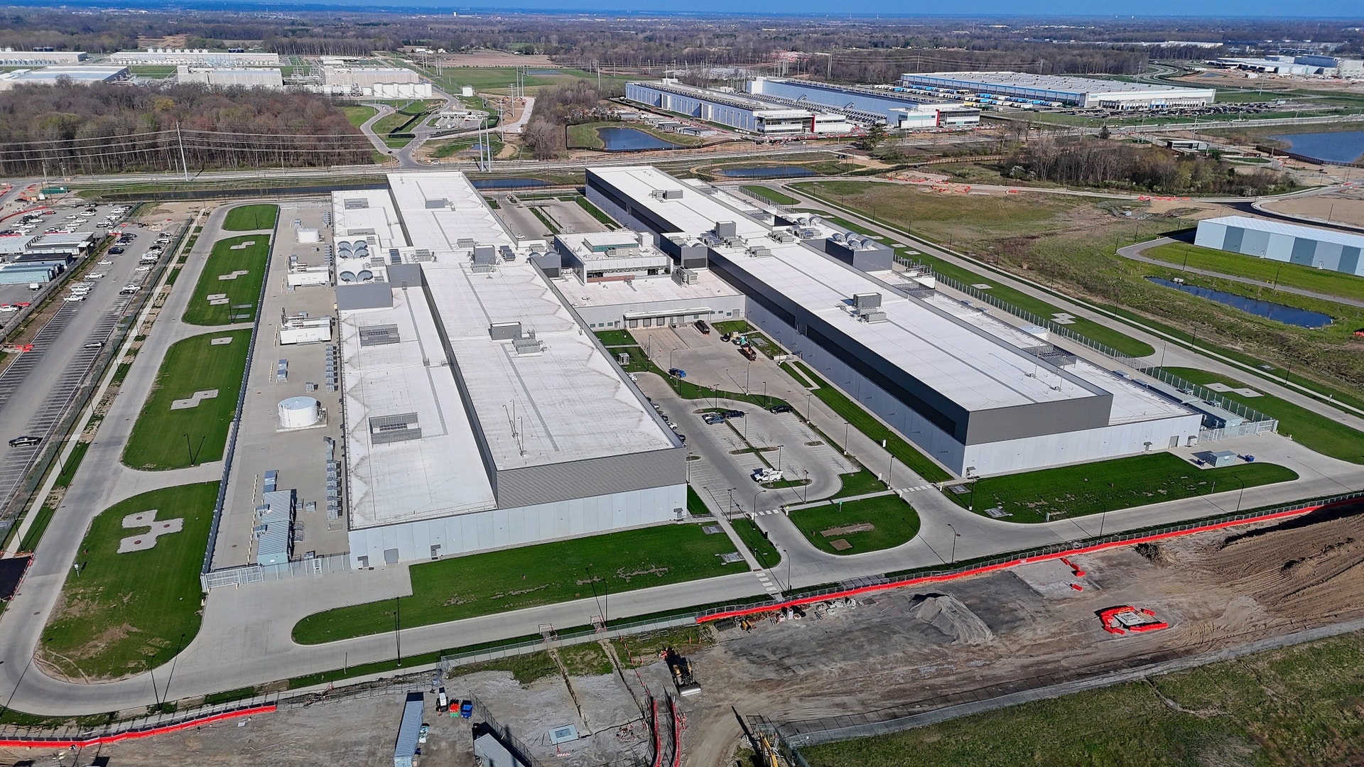 Flygbild över ett stort datacenter med flera ljusa, låga byggnader omgivna av gräsytor, vägar och åkermark.
