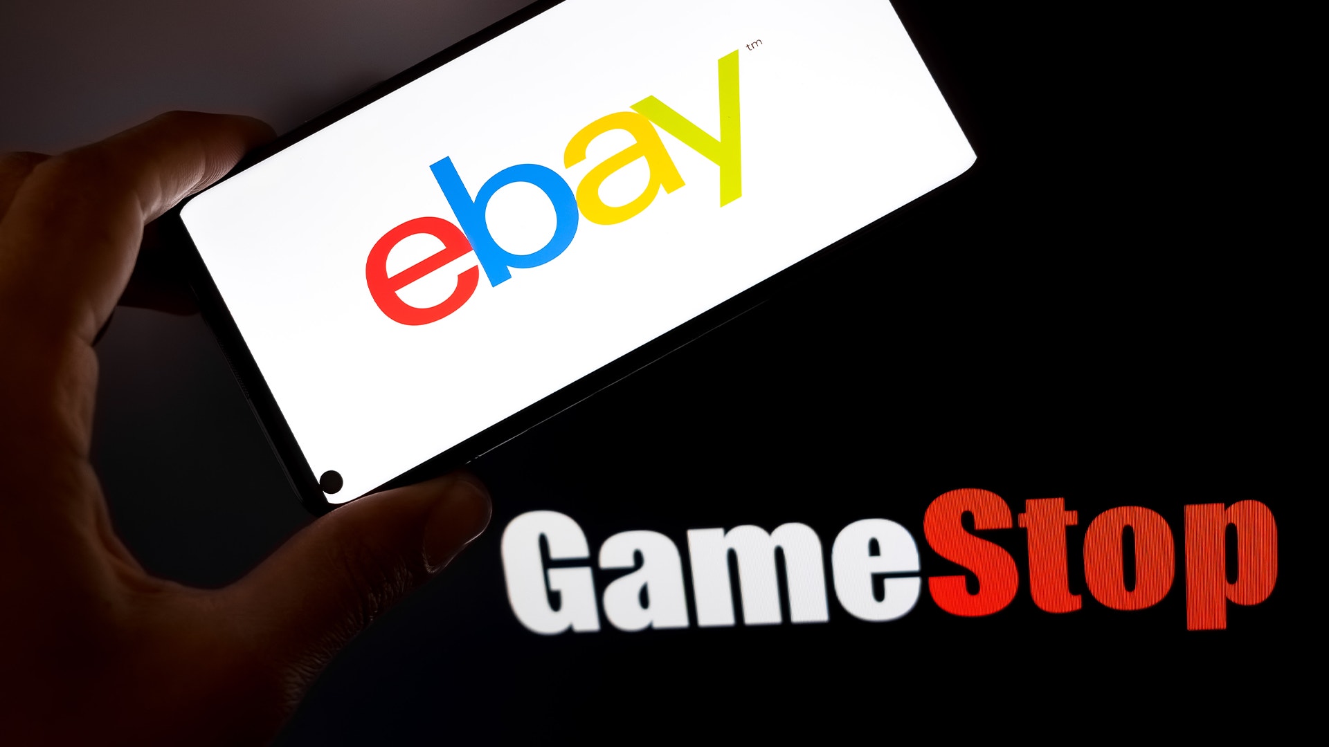 En hand håller en smartphone med eBay-logon. Bakom telefonen syns GameStop-logon på mörk bakgrund.