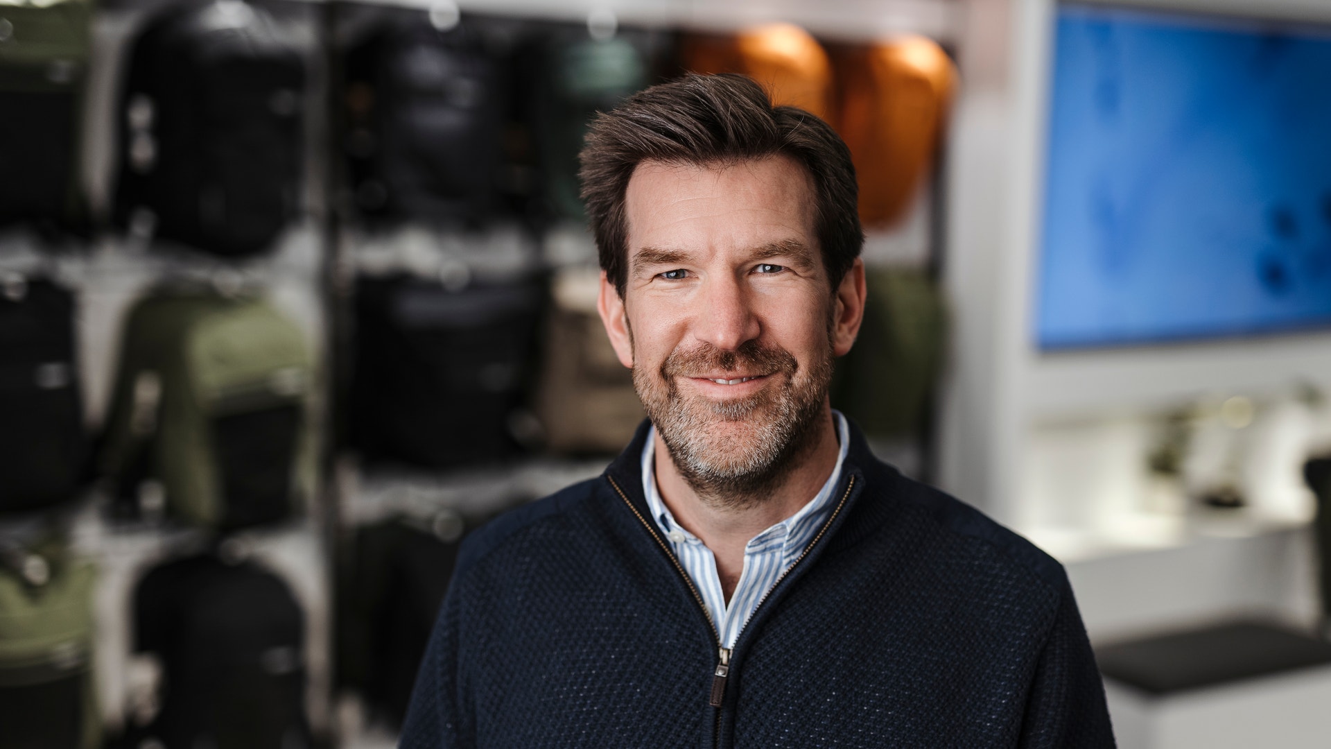 Porträtt av en man i mörkblå tröja framför en suddig bakgrund med industriell utrustning och en blå skärm.