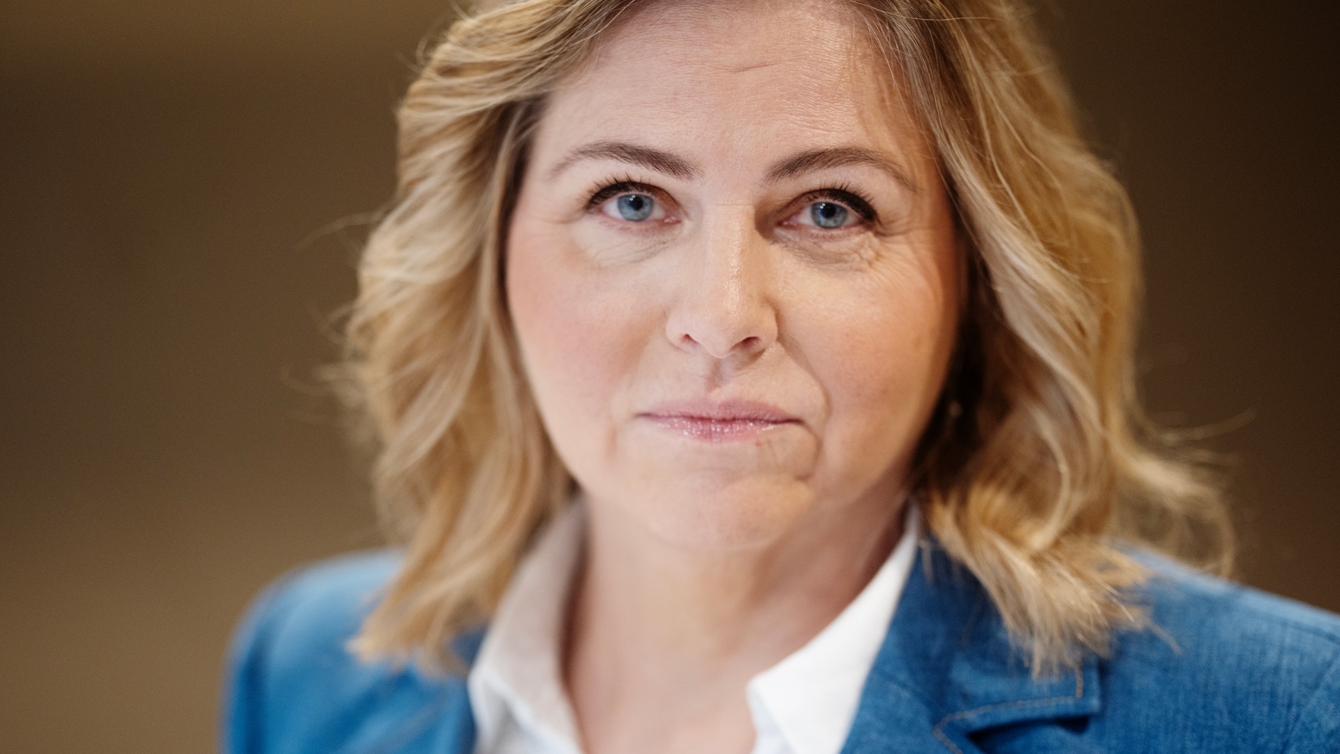 Porträtt av en kvinna med ljust vågigt hår och blå ögon, klädd i blå kavaj och vit skjorta.