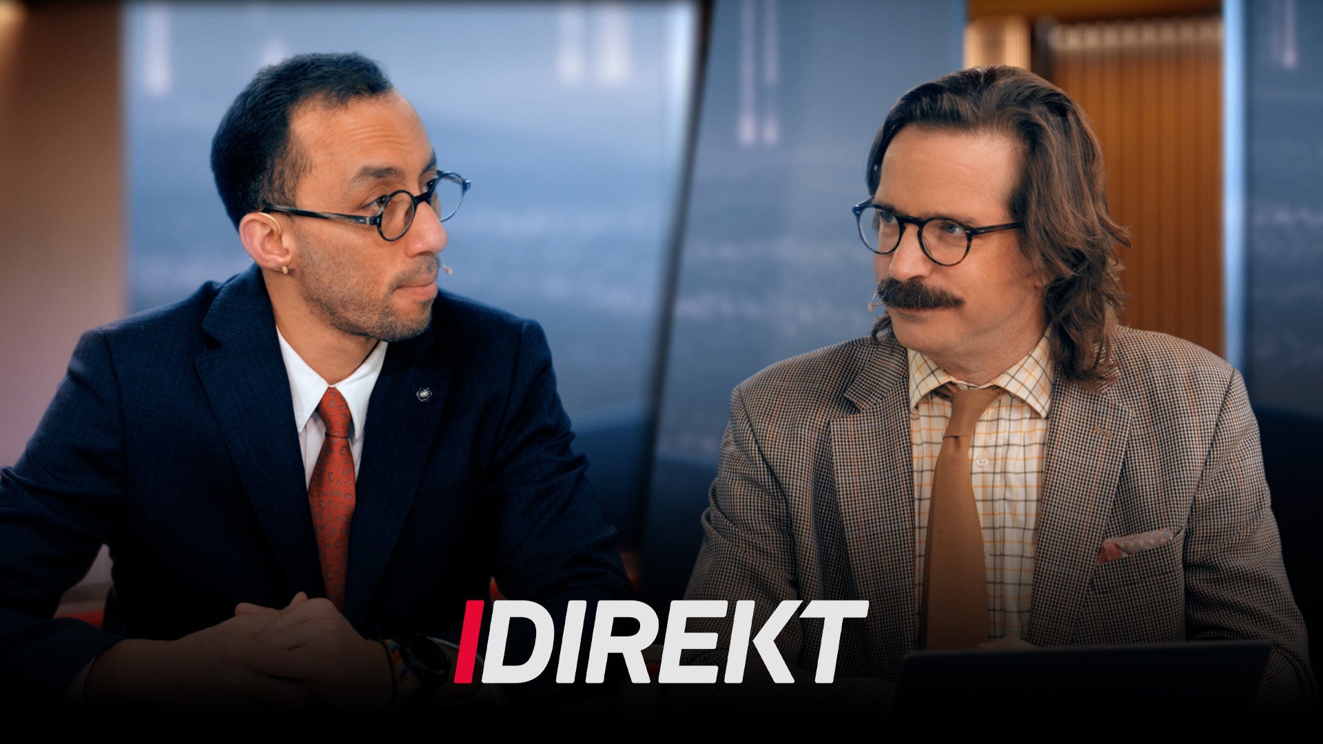 Två män i affärskläder sitter och samtalar i ett kontor med stadsutsikt i bakgrunden.