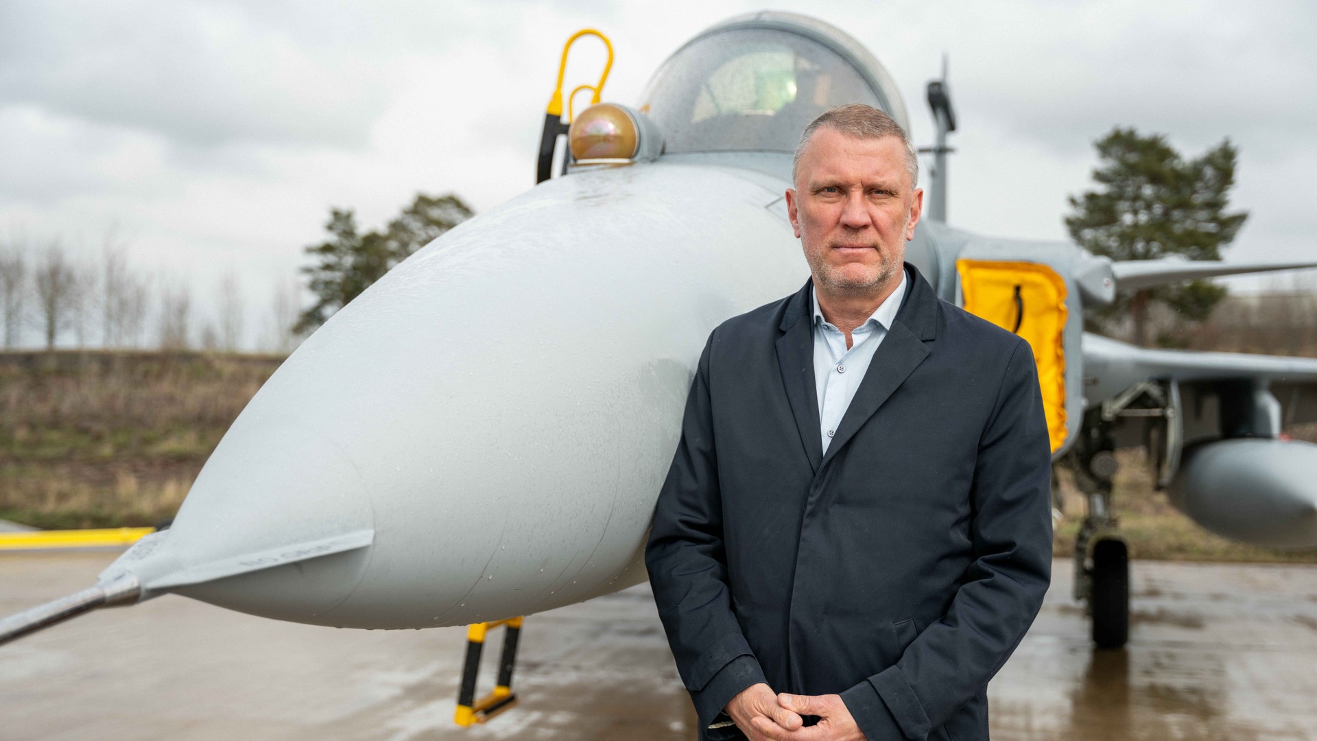 Man i mörk kostym står framför ett militärt stridsflygplan på en flygplats.