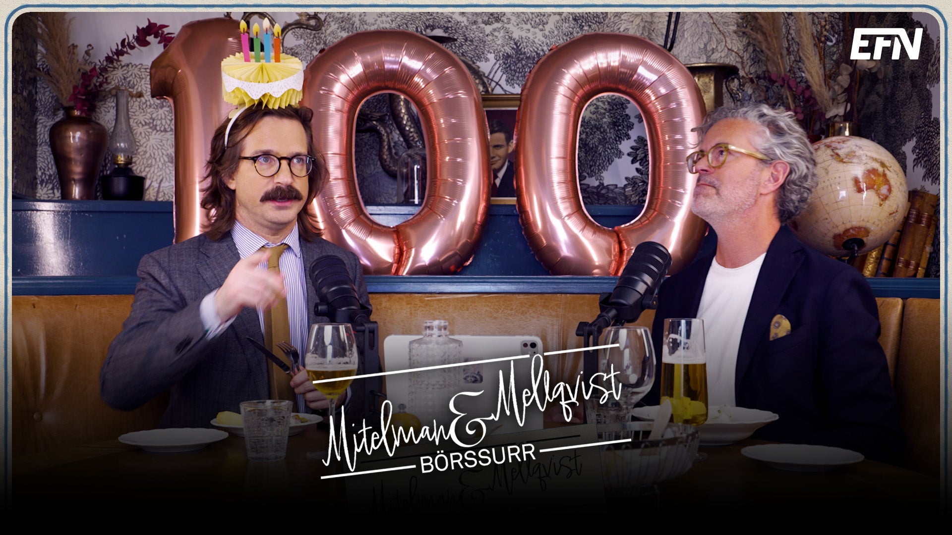 Två män firar vid ett bord med mikrofoner. Bakom dem syns stora röda ballonger med siffran 200. Den ena mannen bär glasögon och en krona.