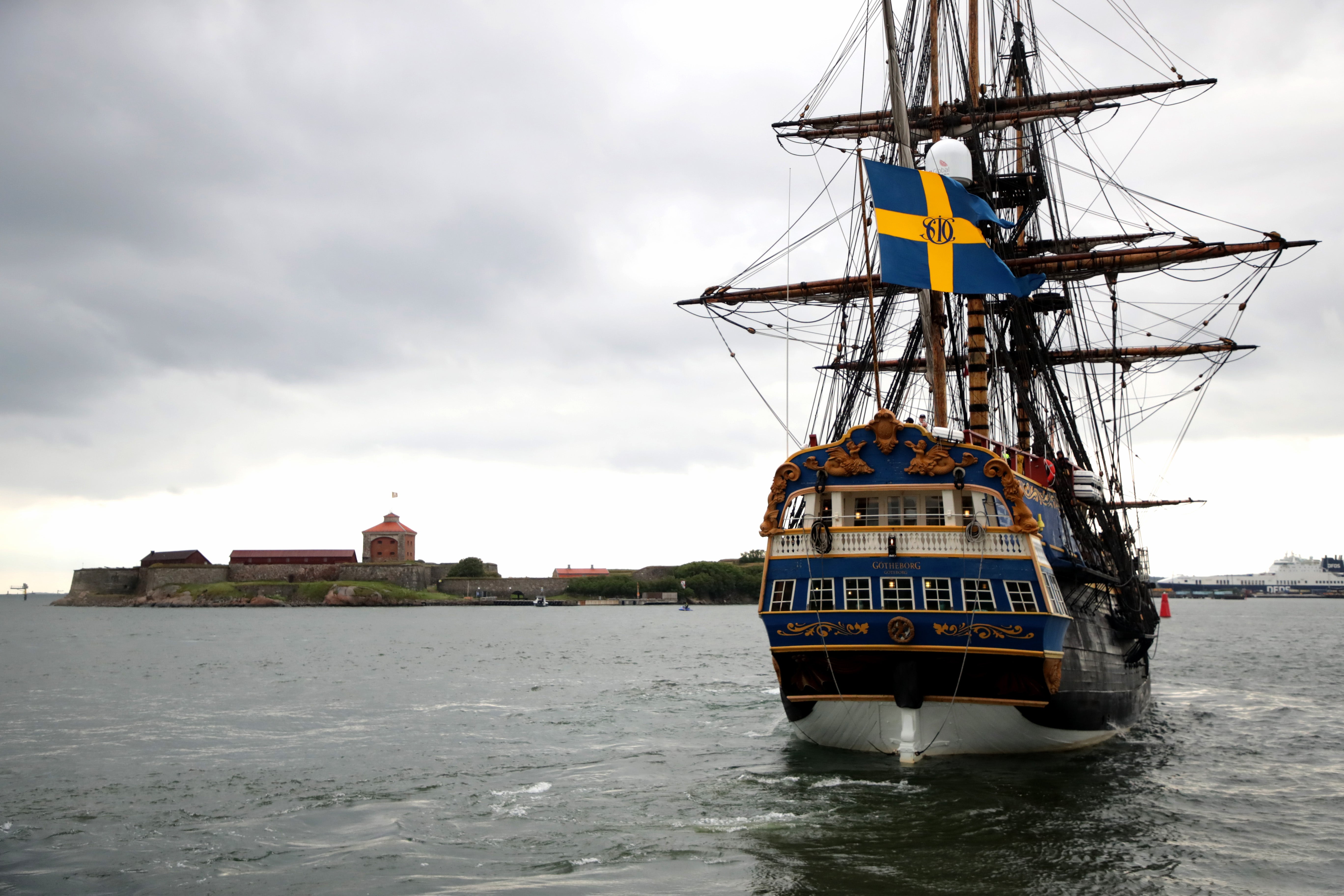 Ett klassiskt segelfartyg med blå och gul skrov och svensk flagga seglar på öppet vatten. En befäst ö syns i bakgrunden.