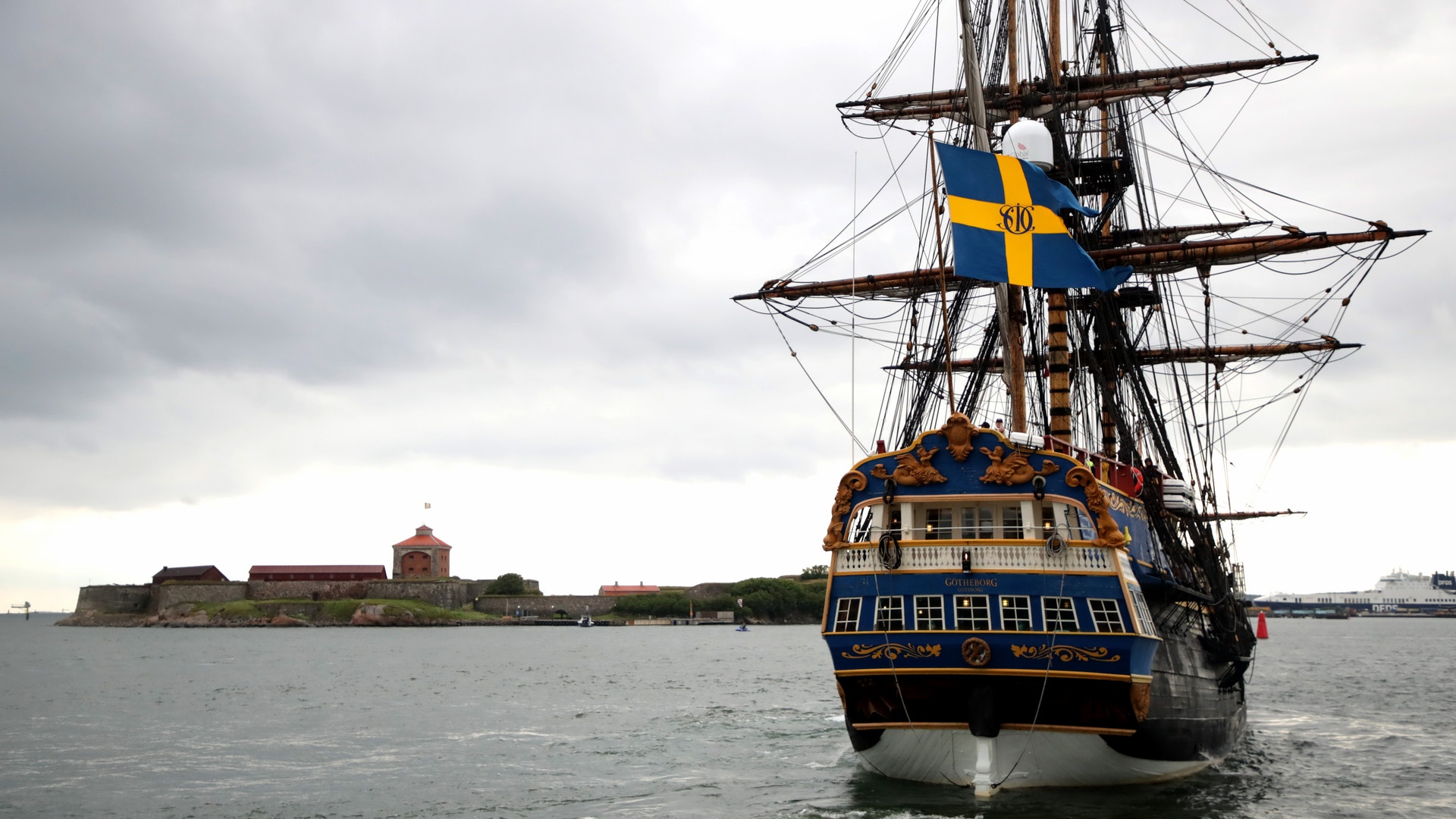 Ett klassiskt segelfartyg med blå och gul skrov och svensk flagga seglar på öppet vatten. En befäst ö syns i bakgrunden.