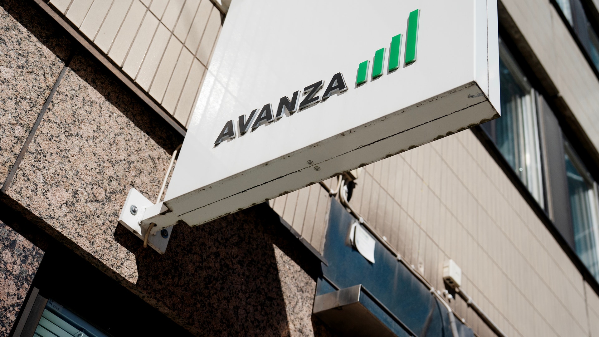 Vit skylt med texten Avanza och en grön stapeldiagram-logotyp monterad på en byggnadsfasad.