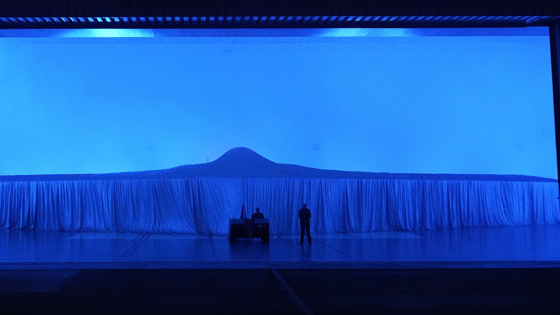 En teatralisk scen med blå belysning där en ensam person står framför ett stort blått gardinöppet med ett berg synligt i bakgrunden.