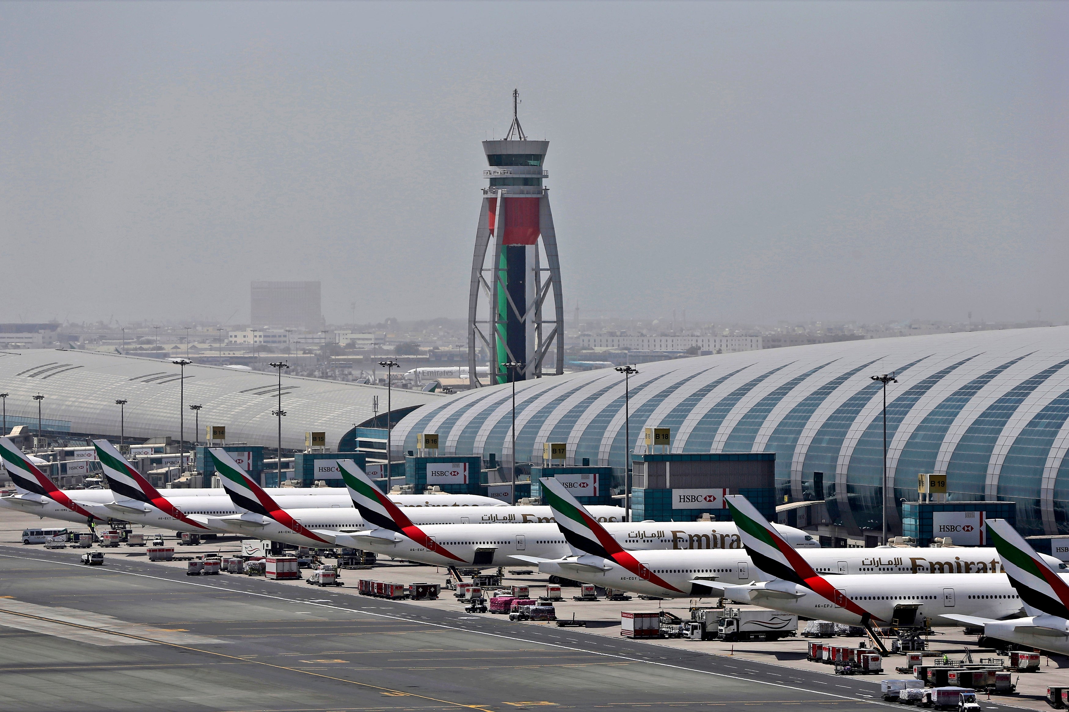 Flygplats med flera vita Emirates-flygplan parkerade vid terminalbyggnaden. En kontrolltourn med UAE:s flaggfärger syns i bakgrunden tillsammans med stadshorisont.
