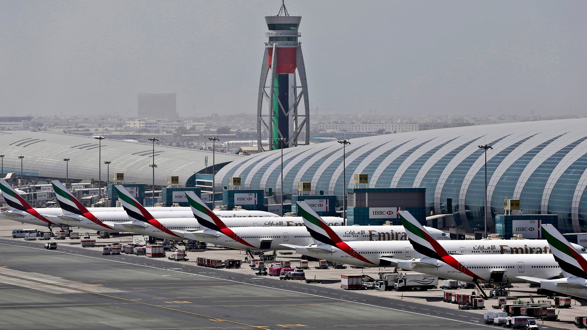 Flygplats med flera vita Emirates-flygplan parkerade vid terminalbyggnaden. En kontrolltourn med UAE:s flaggfärger syns i bakgrunden tillsammans med stadshorisont.