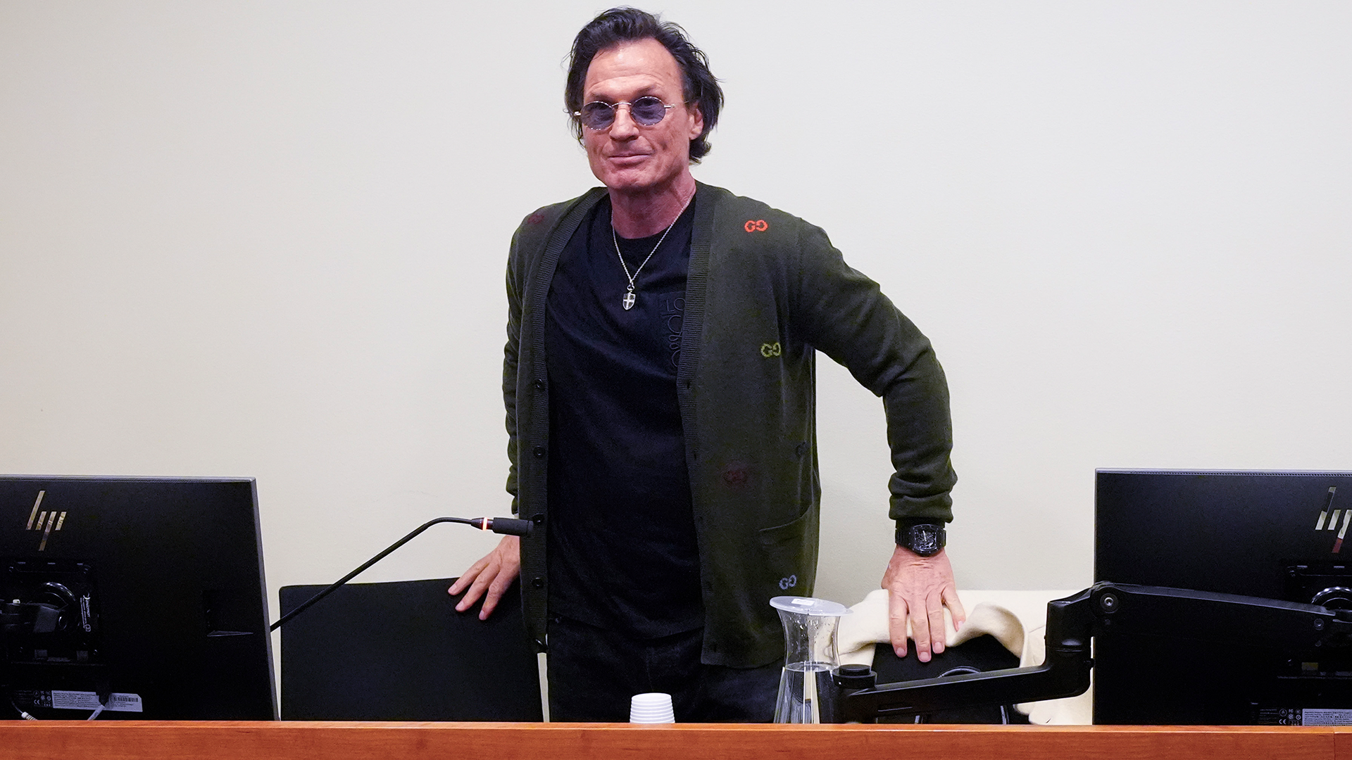 Petter Stordalen, i glasögon och mörk jacka, står vid ett skrivbord med datorskärmar.