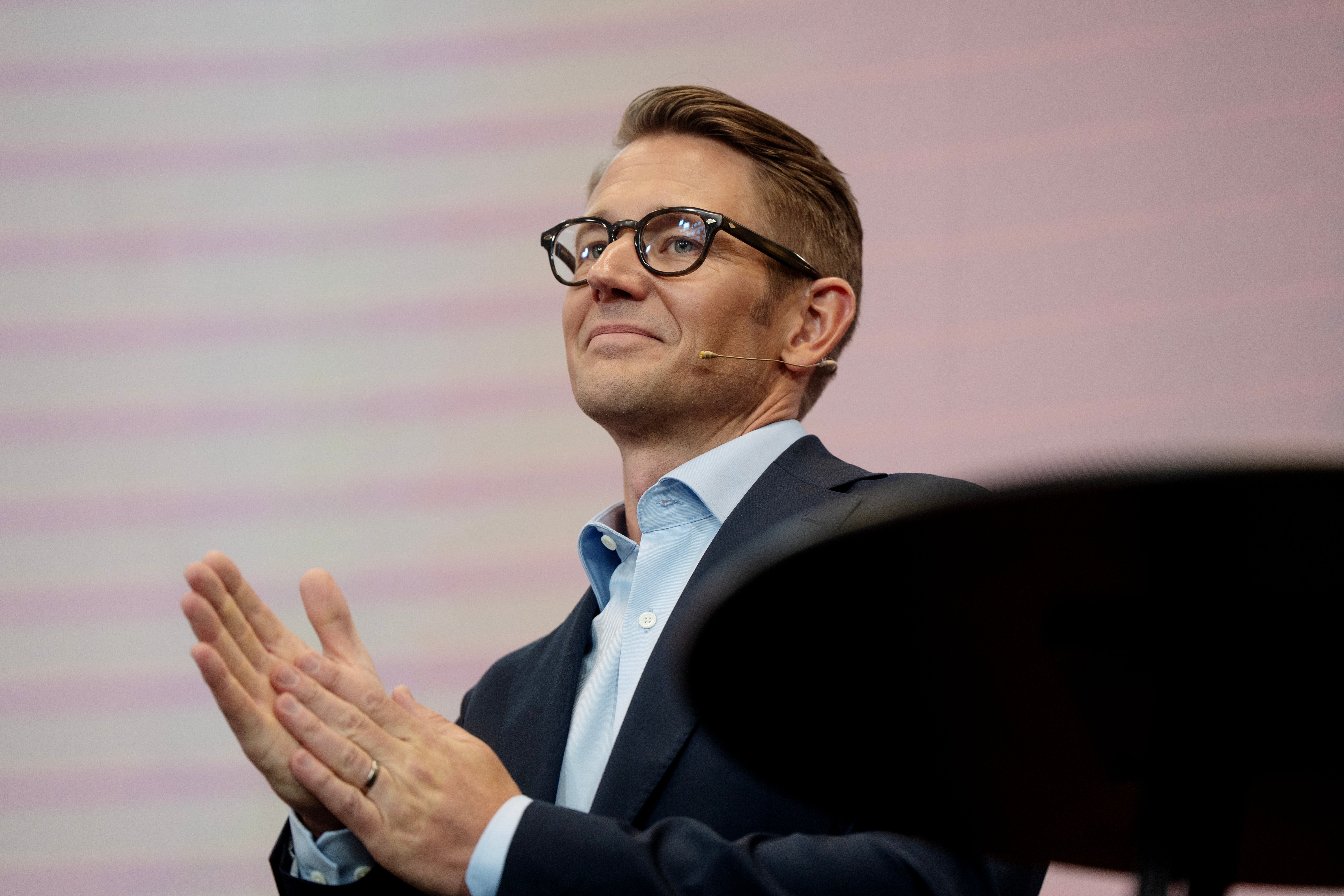 Man med glasögon i mörkblå kavaj och ljusblå skjorta håller en presentation och gestikulerar med händerna. Rosa bakgrund med horisontella streck.