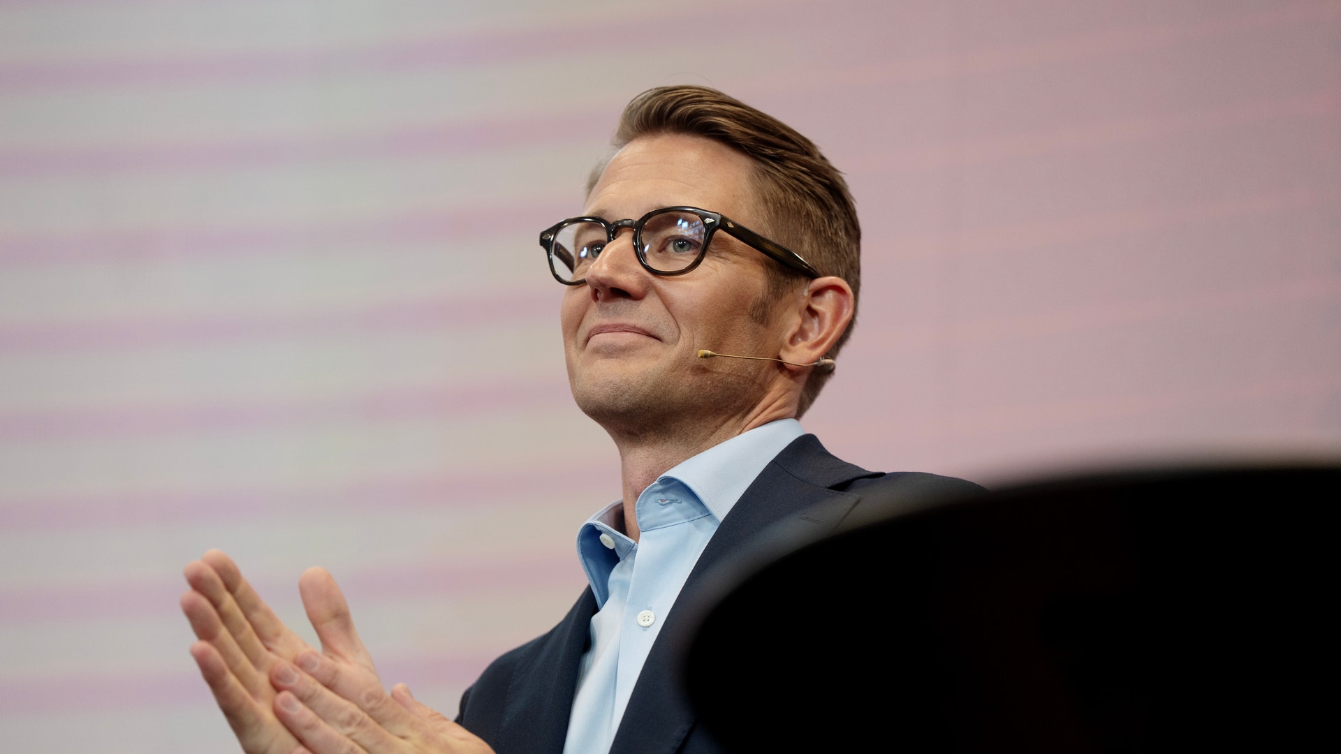 Man med glasögon i mörkblå kavaj och ljusblå skjorta håller en presentation och gestikulerar med händerna. Rosa bakgrund med horisontella streck.