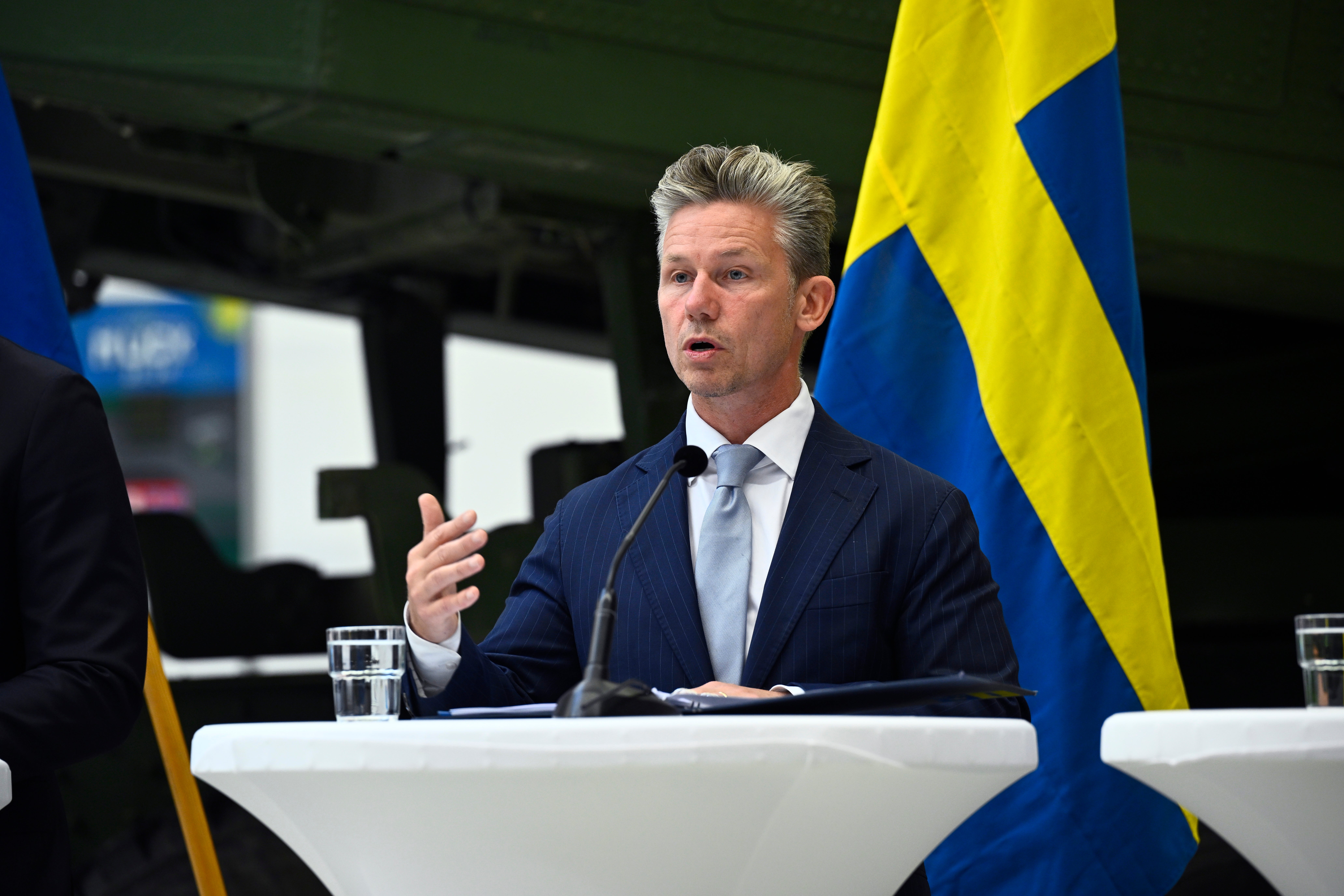 Man i mörkblå kostym talar vid ett podium med svenska flaggor i bakgrunden.