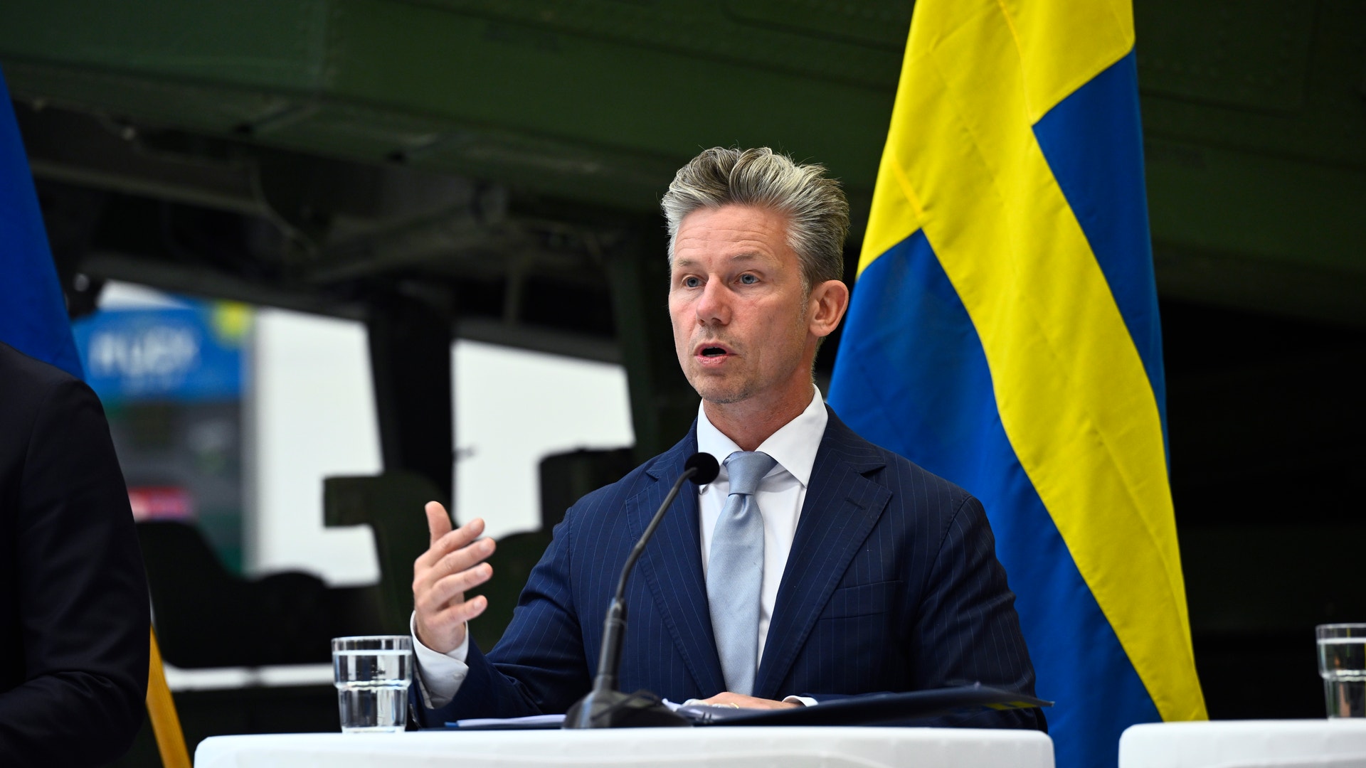 Man i mörkblå kostym talar vid ett podium med svenska flaggor i bakgrunden.