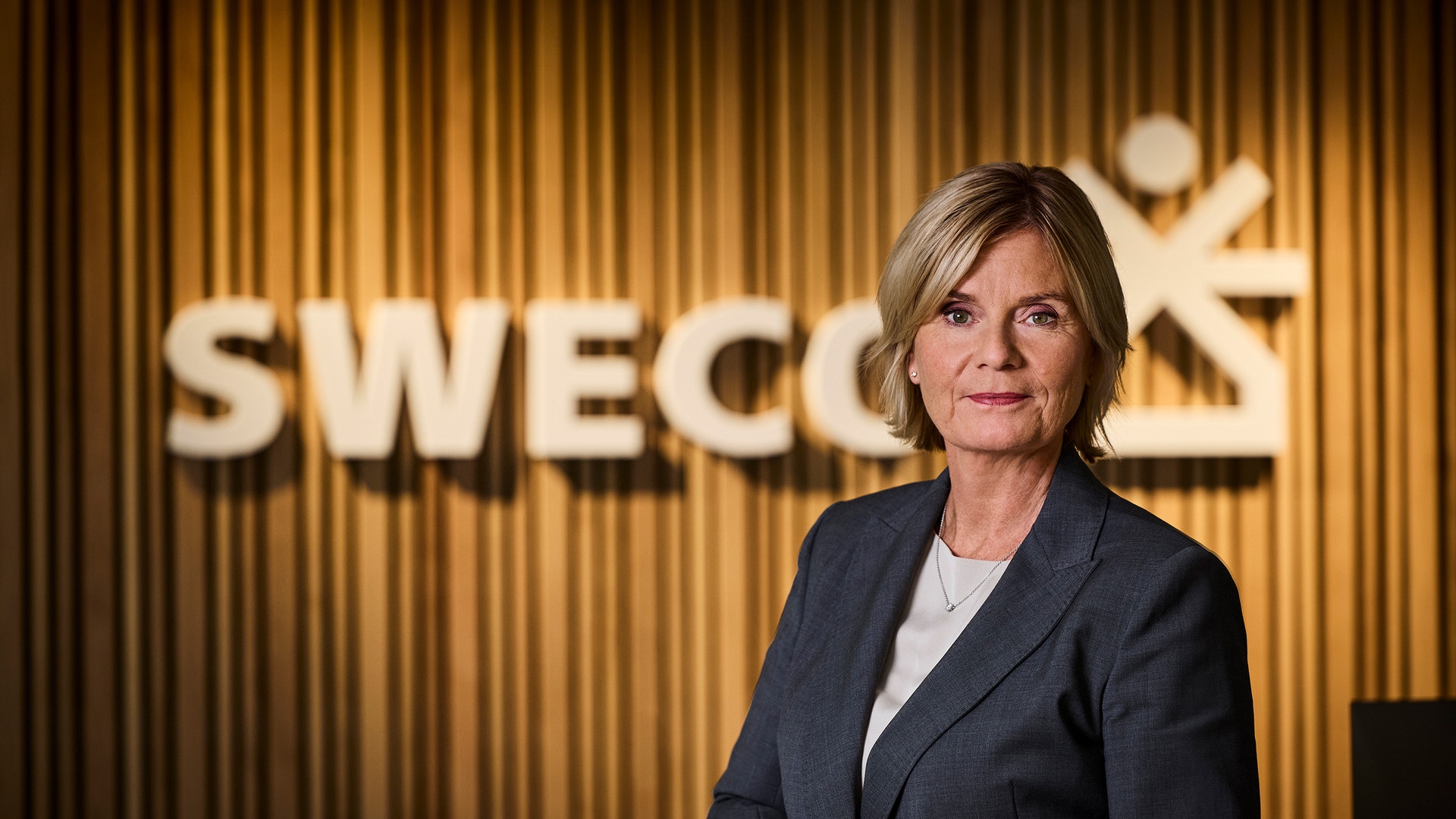 Porträtt av en kvinna i mörkblå kavaj framför Swecos gula vägg med logotyp.