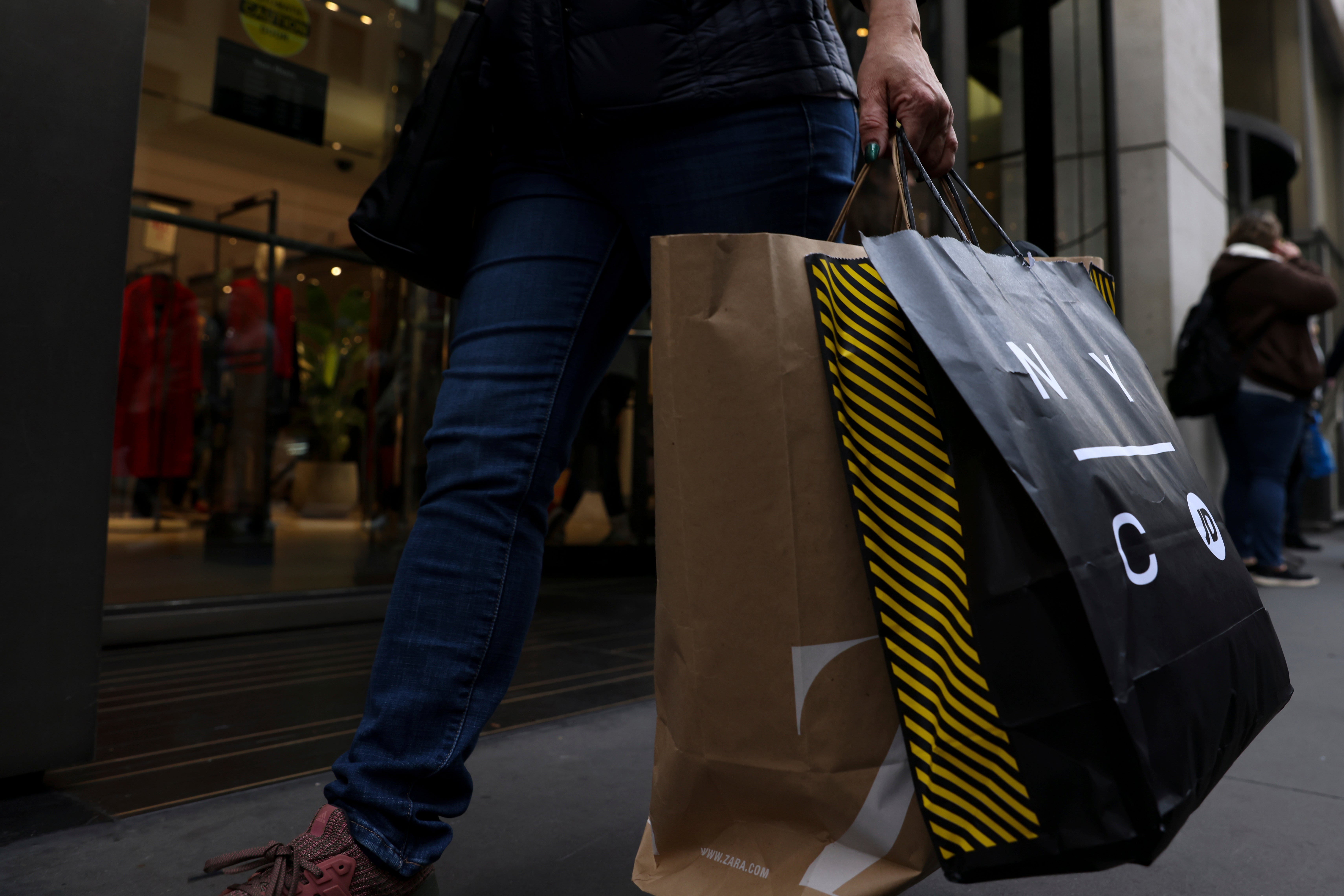 Person i blå jeans som bär shoppingväskor framför ett butiksfönster.