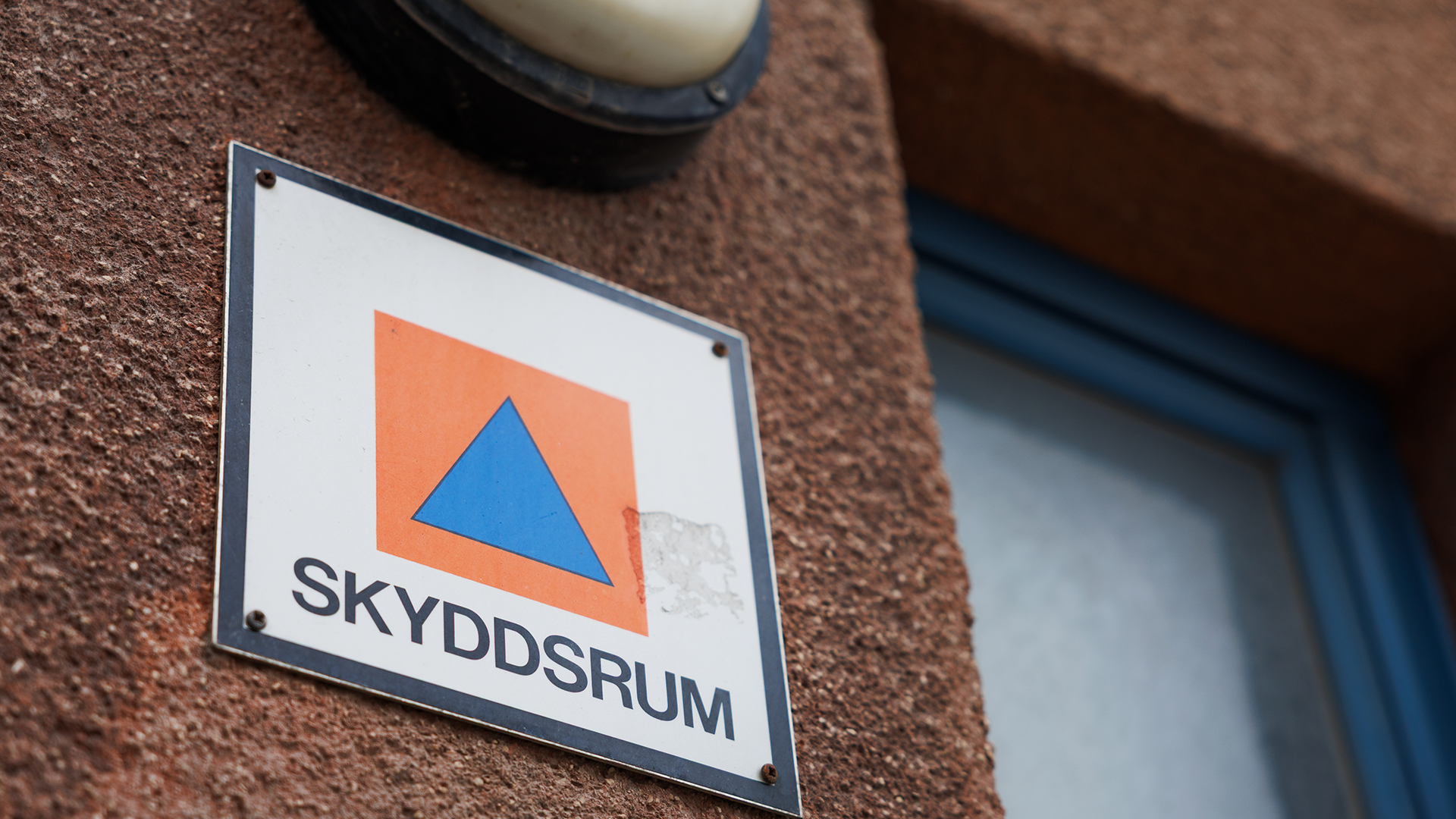 Skylt på tegelväggen med texten SKYDDSRUM och en symbol bestående av en blå triangel på orange bakgrund.