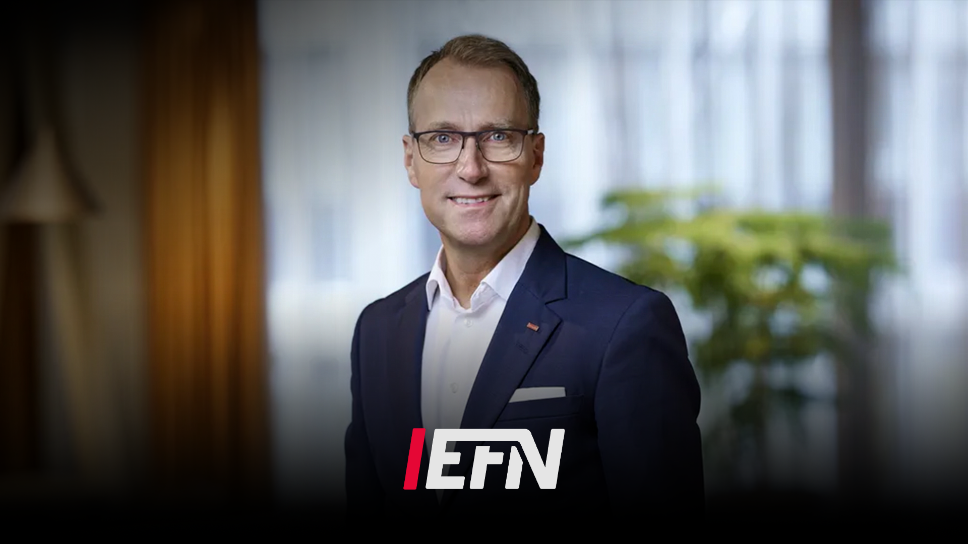 Porträtt av en man i mörk kostym och glasögon som ler mot kameran i en modern kontorsmiljö. EFN-logon är synlig i bildens nedre del.