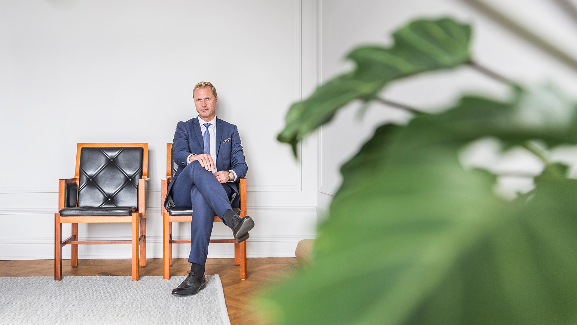 Man i blå kostym sitter på en stol i ett ljust kontor omgivet av gröna växter.