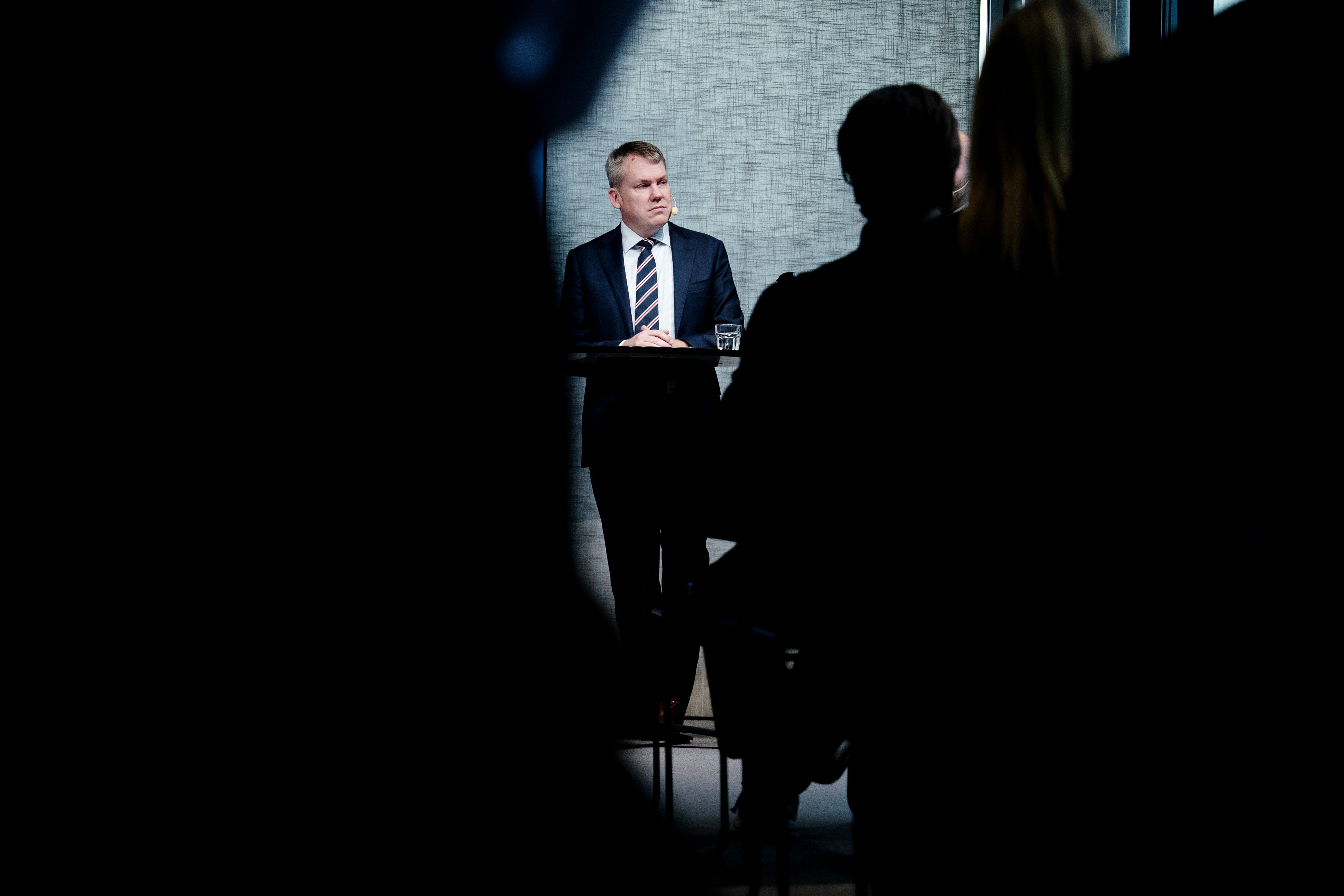 En man i kostym och slips står vid ett litet högt bord och blickar åt sidan. I förgrunden syns mörka siluetter av publiken.