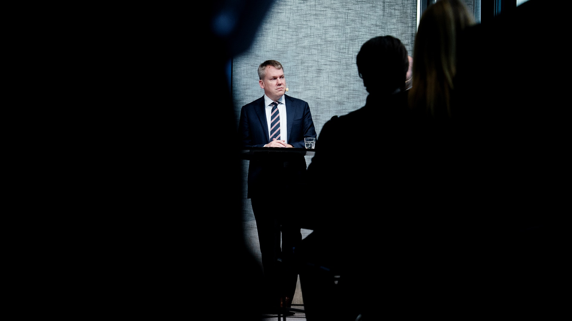 En man i kostym och slips står vid ett litet högt bord och blickar åt sidan. I förgrunden syns mörka siluetter av publiken.
