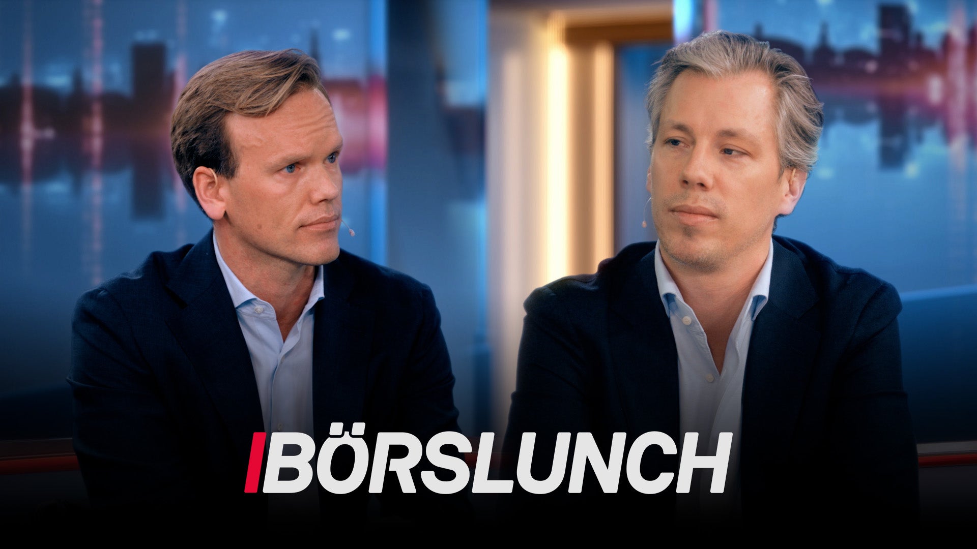 Två män i affärskläder diskuterar i en TV-studio med stadshorisont i bakgrunden.