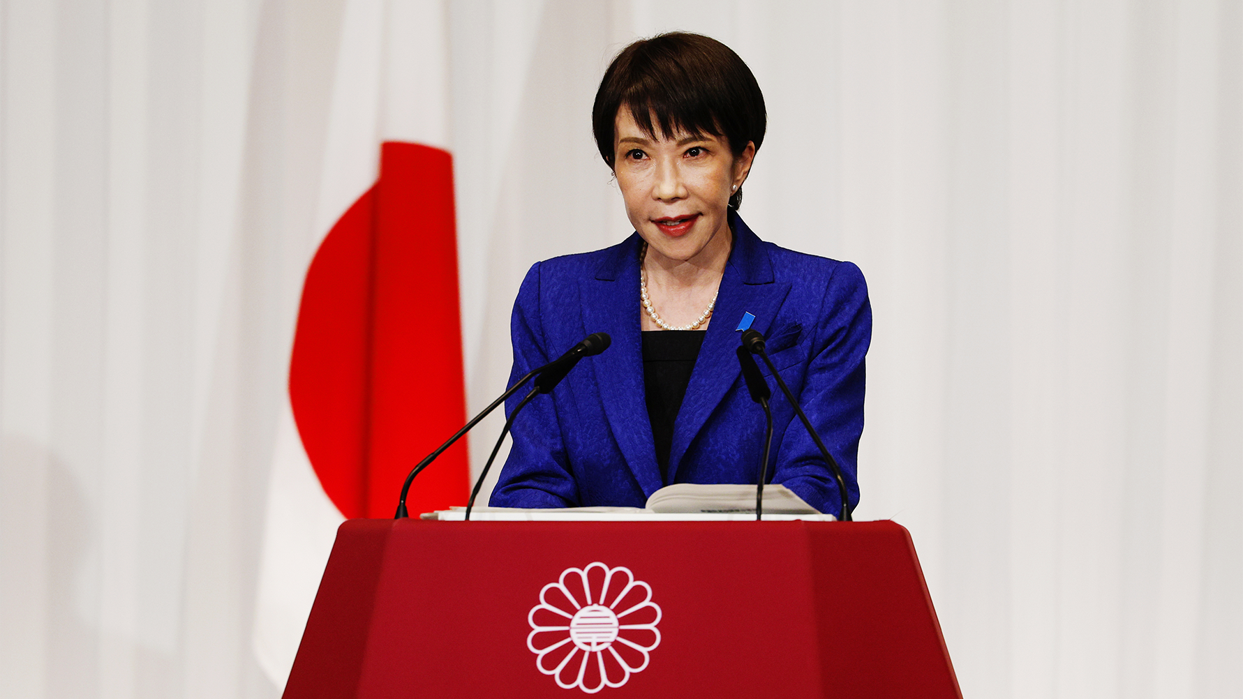 Kvinna i blå kavaj talar vid ett podium med Japans flagga i bakgrunden