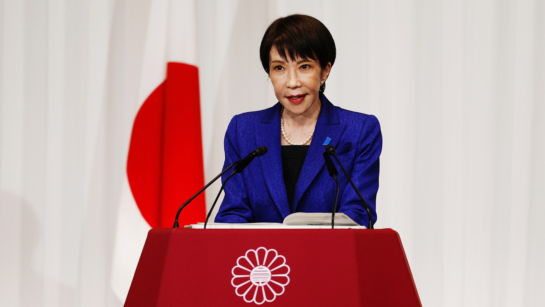 Kvinna i blå kavaj talar vid ett podium med Japans flagga i bakgrunden