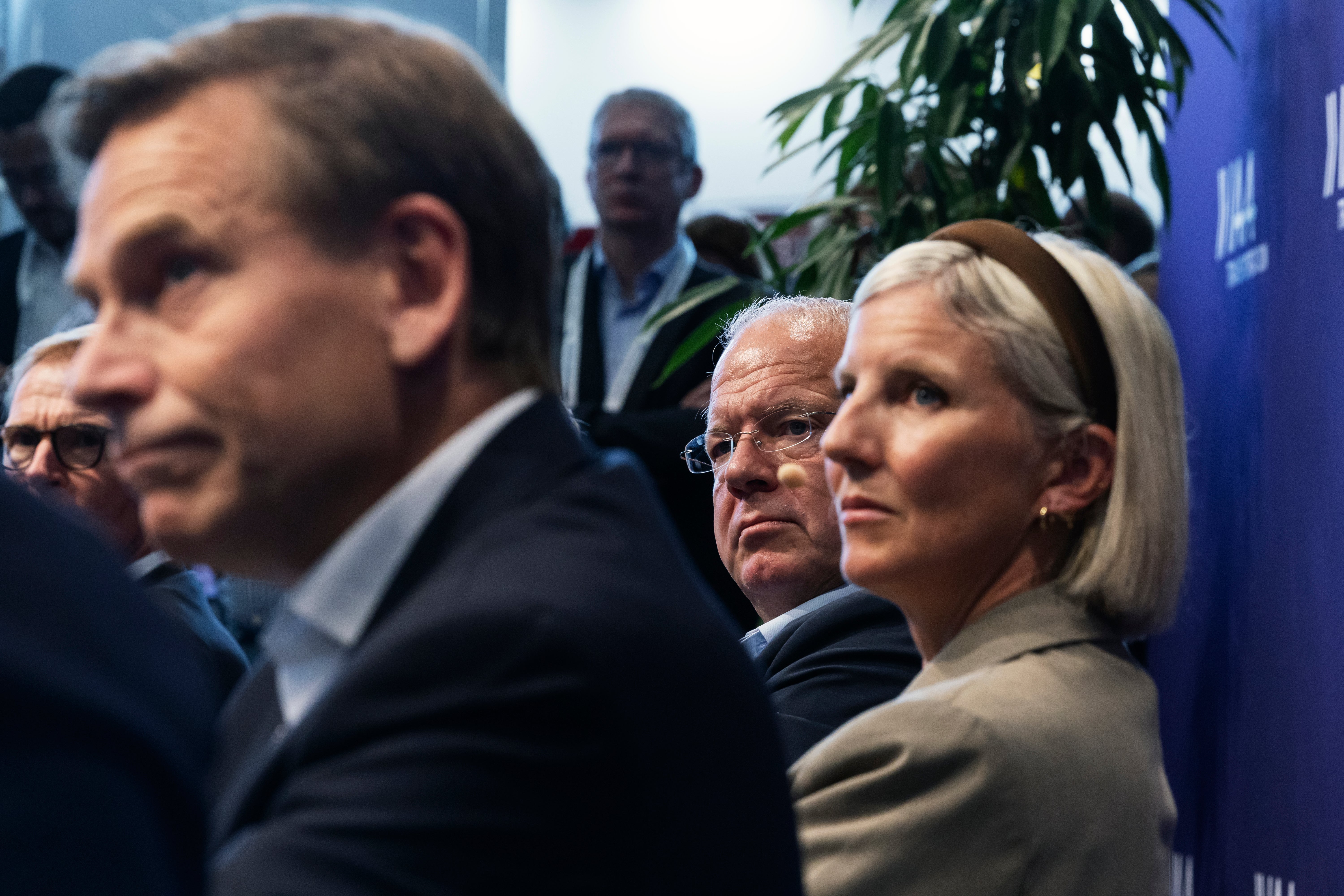 En grupp män och kvinnor i kostym lyssnar uppmärksamt vid ett evenemang inomhus.