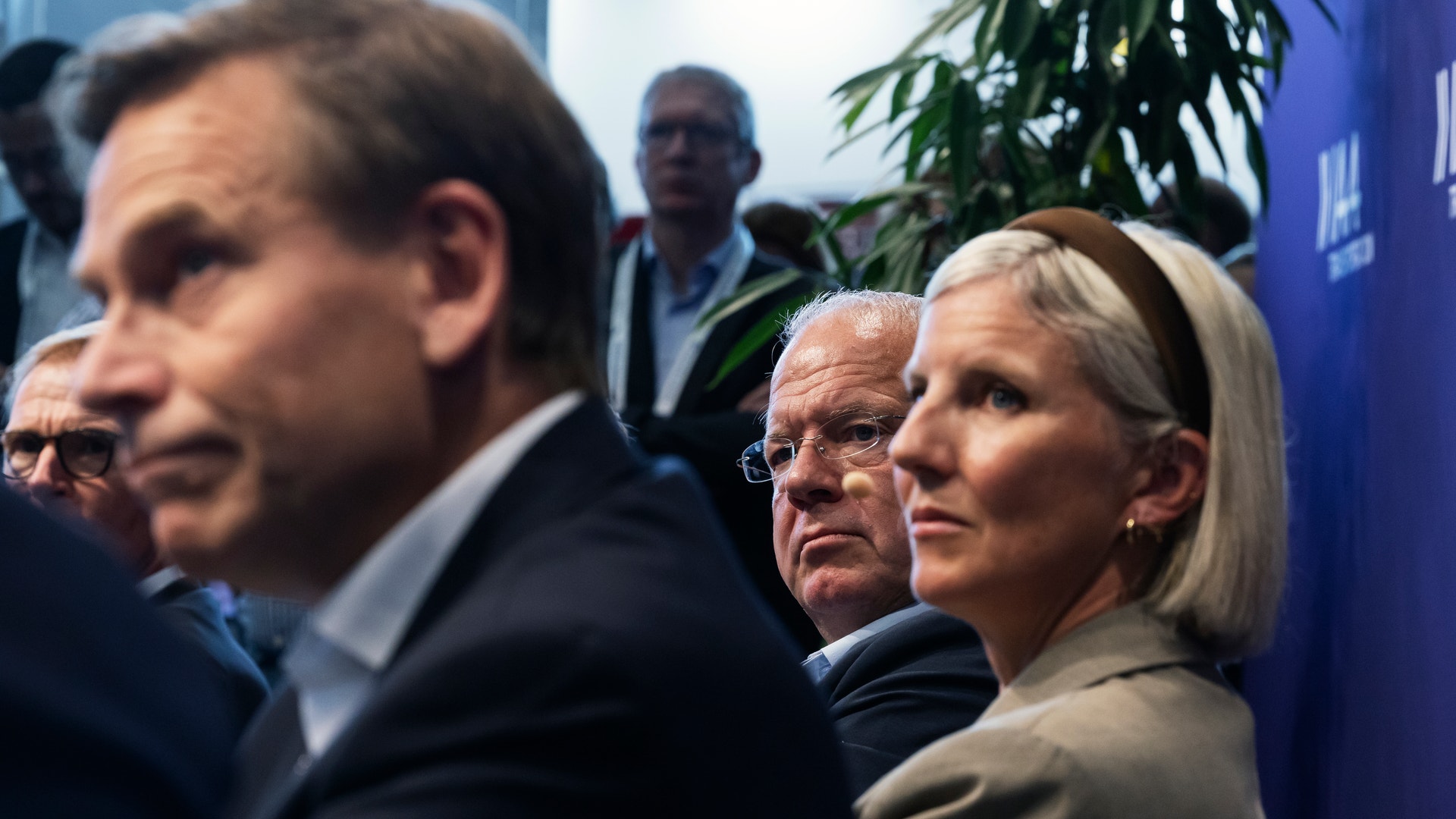 En grupp män och kvinnor i kostym lyssnar uppmärksamt vid ett evenemang inomhus.