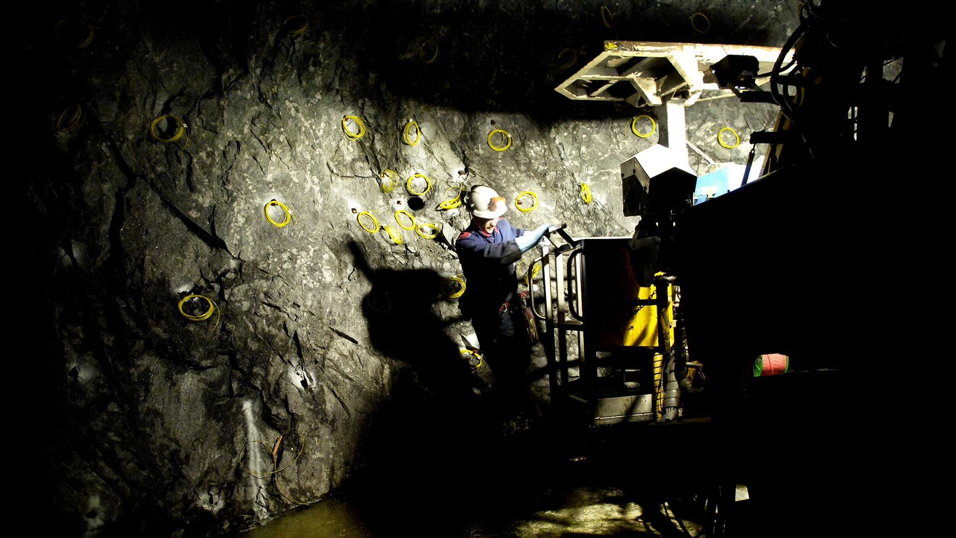 En gruvarbetare i skyddsutrustning och hjälm med pannlampa arbetar vid en bergvägg i en underjordisk gruvschakt. Bergväggen är markerad med gula cirklar och belyst av industriell belysning. Gul gruvutrustning och säkerhetsutrustning syns i bakgrunden.