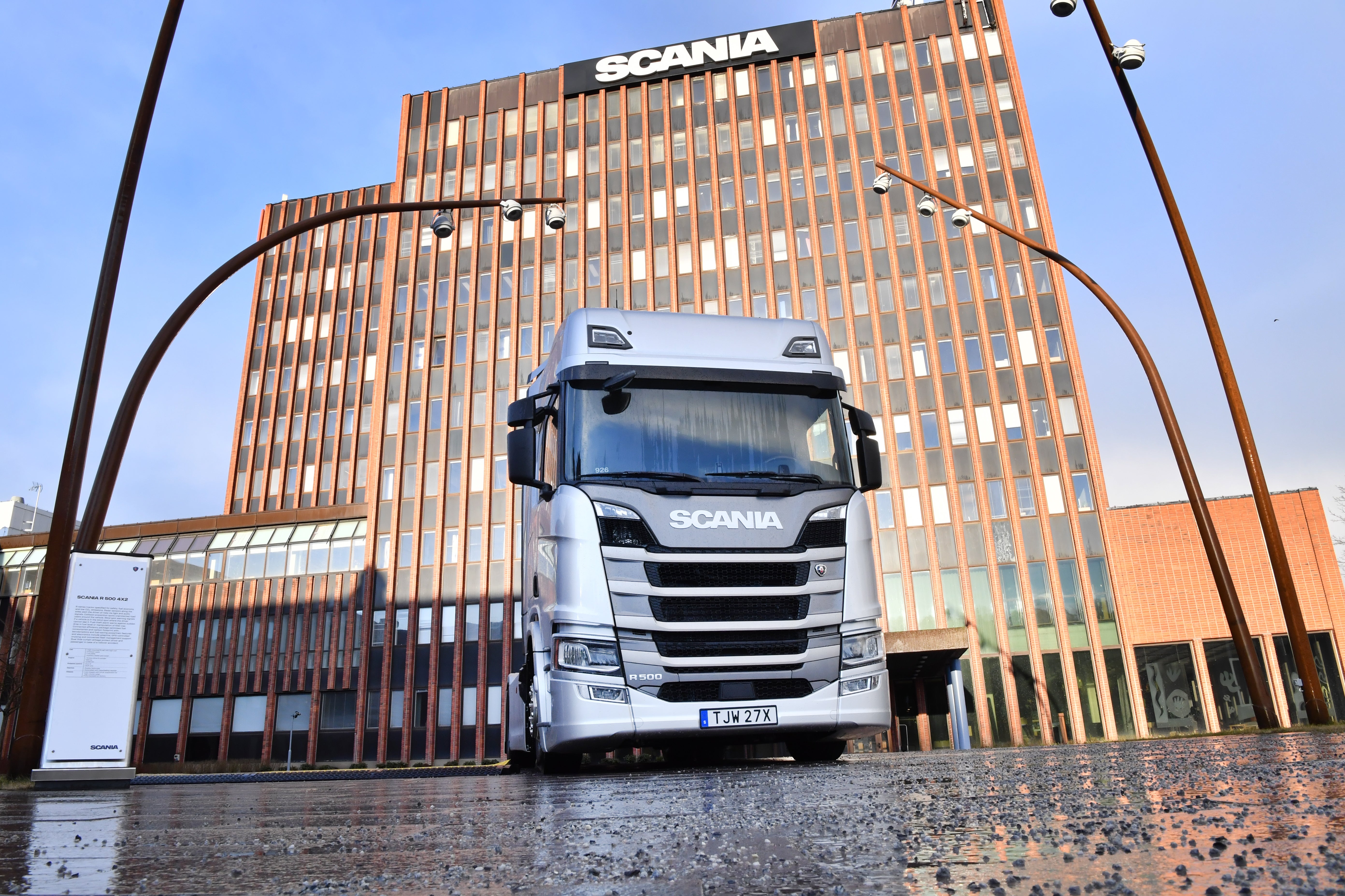 En vit Scania-lastbil står framför Scanias huvudkontor, en stor modern byggnad med orange och bruna fasader.