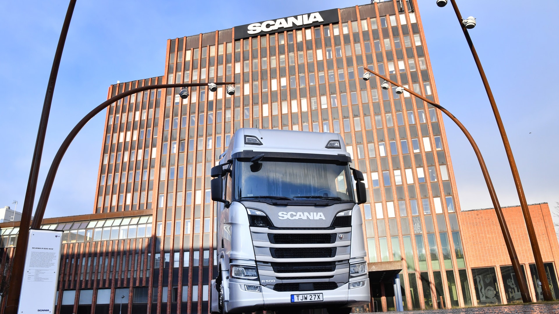 En vit Scania-lastbil står framför Scanias huvudkontor, en stor modern byggnad med orange och bruna fasader.