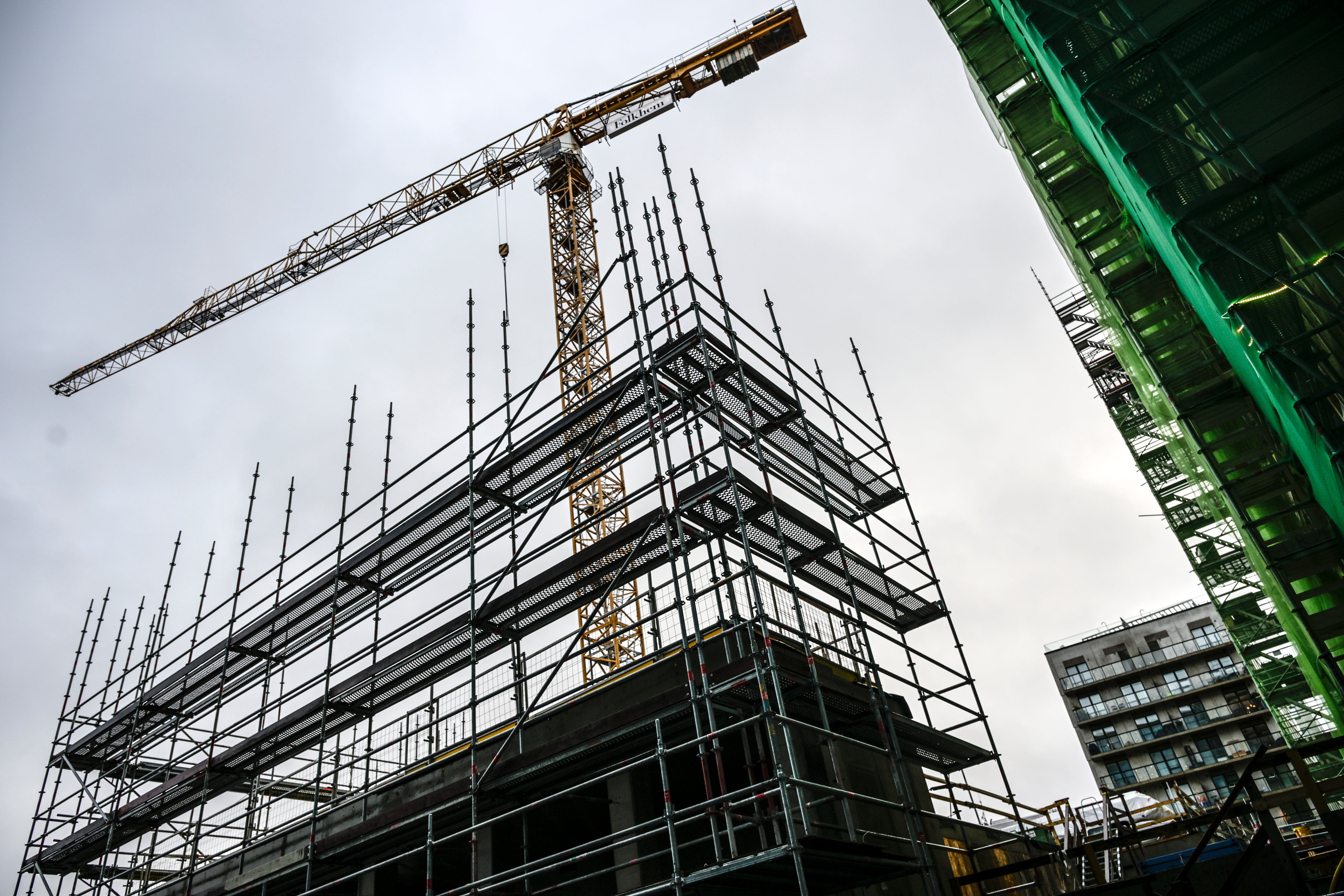 En byggarbetsplats med ställningar och stål-konstruktion under uppförande. En stor gul byggkran är monterad ovanför byggnaden. Moderna bostadshus syns i bakgrunden.