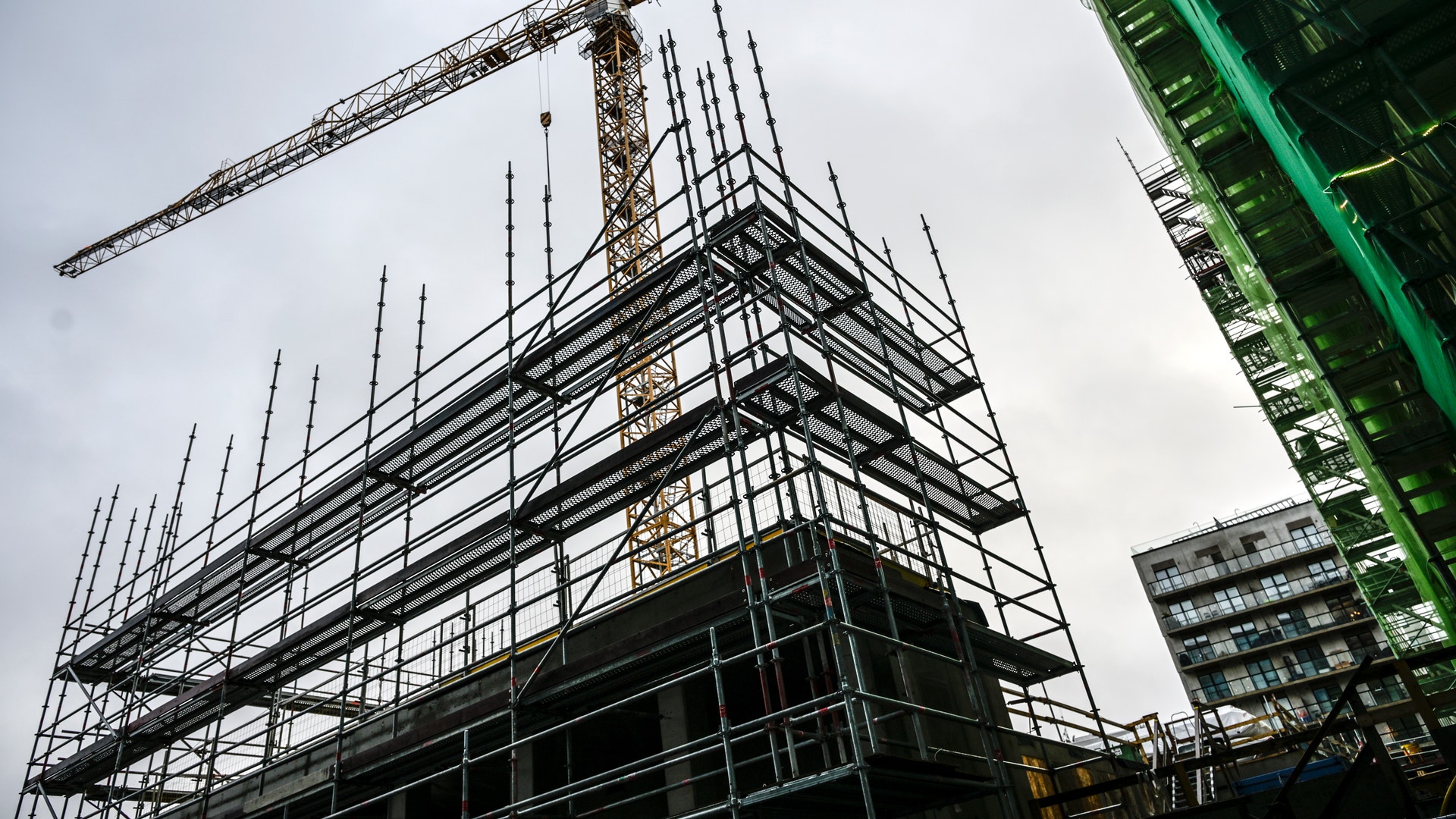 En byggarbetsplats med ställningar och stål-konstruktion under uppförande. En stor gul byggkran är monterad ovanför byggnaden. Moderna bostadshus syns i bakgrunden.